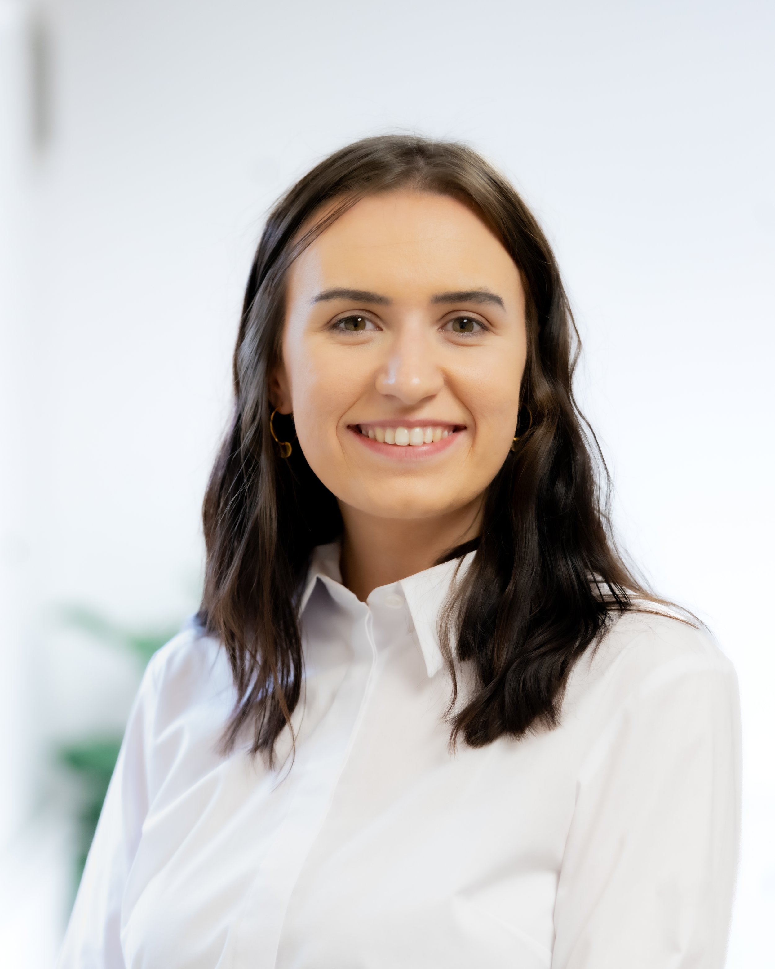 A woman with shoulder-length dark brown hair wearing a white button-up shirt, smiling, with a blurred indoor background.