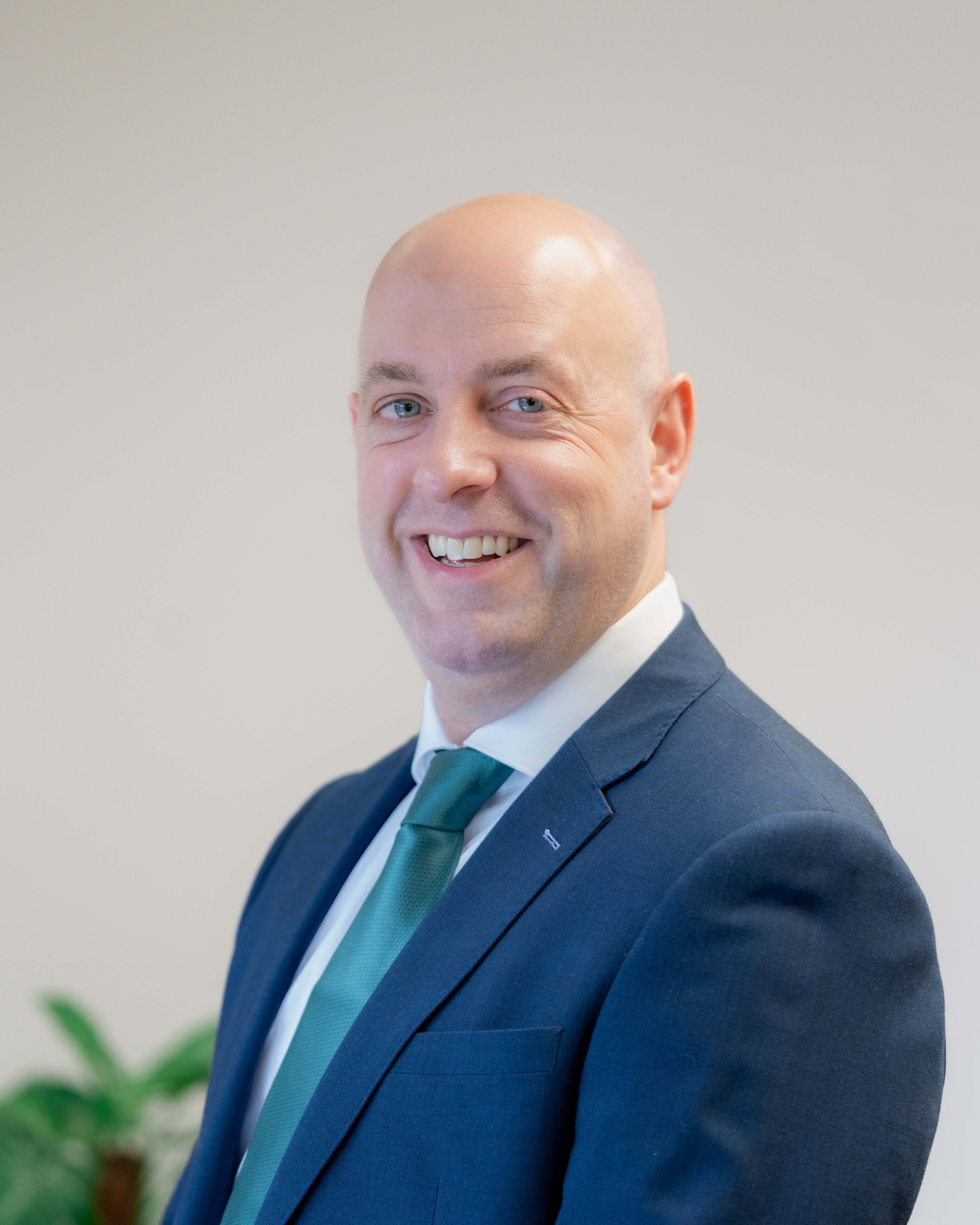 Professional portrait of a smiling bald man in a navy suit with a green tie, standing against a plain background.