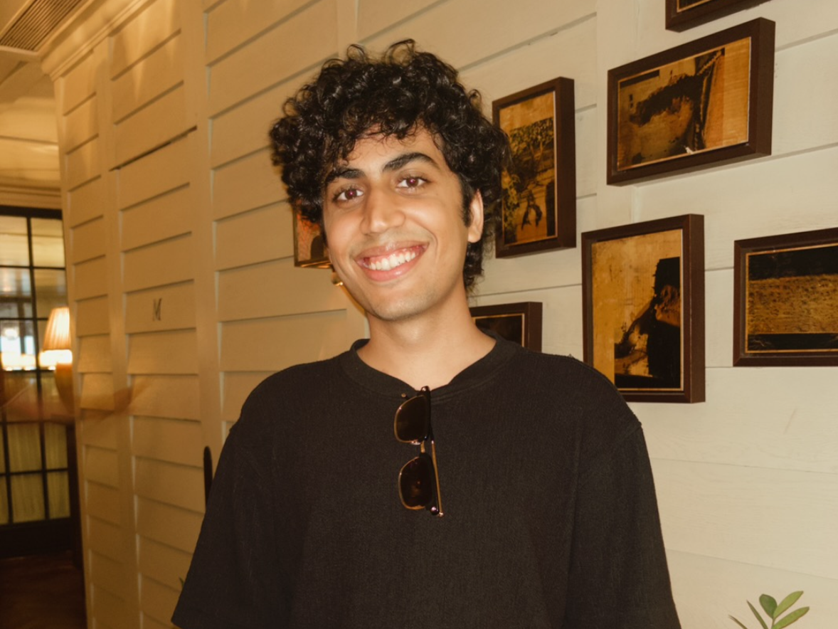 Smiling young man with curly hair wearing a black shirt and sunglasses hanging on his collar, standing in a warmly lit room with framed photos on the wall behind him.