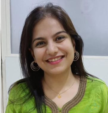 A woman with long dark hair, wearing a green top with gold embellishments, smiling in front of a white wall.