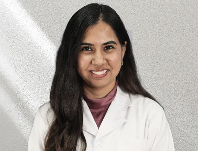 A young woman with long dark hair, wearing a white lab coat and a pink turtleneck, smiling at the camera against a grey textured wall background.