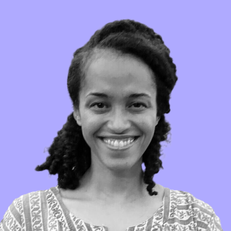 A woman with curly hair styled in an asymmetrical cut, smiling and looking directly at the camera, wearing a patterned top, against a purple background.