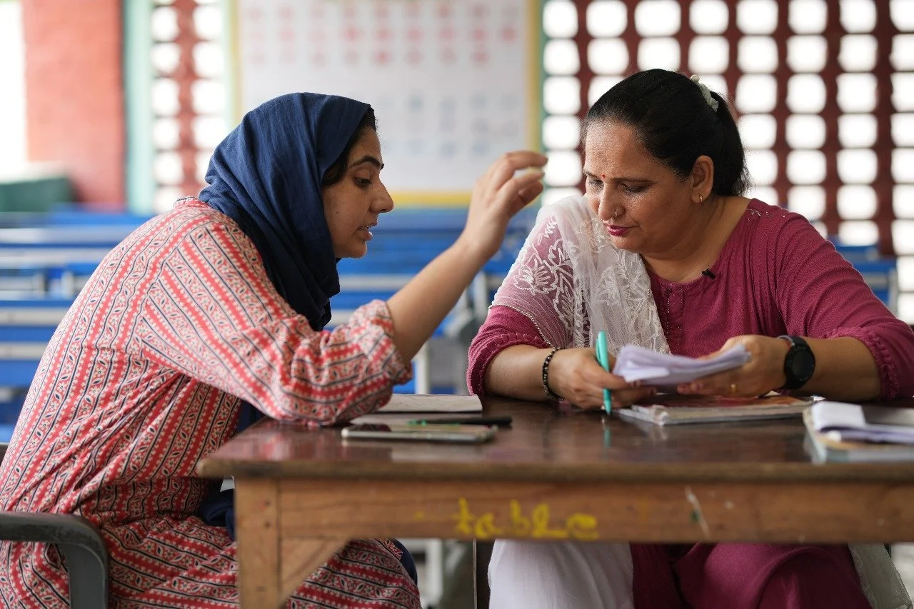 Two women are sitting at a wooden desk in a classroom, engaged in a discussion over papers and notebooks. One woman, wearing a blue headscarf and a patterned dress, is speaking and gesturing with her hand, while the other woman, wearing a maroon outfit with a white shawl, is listening attentively while holding a pen and some papers.