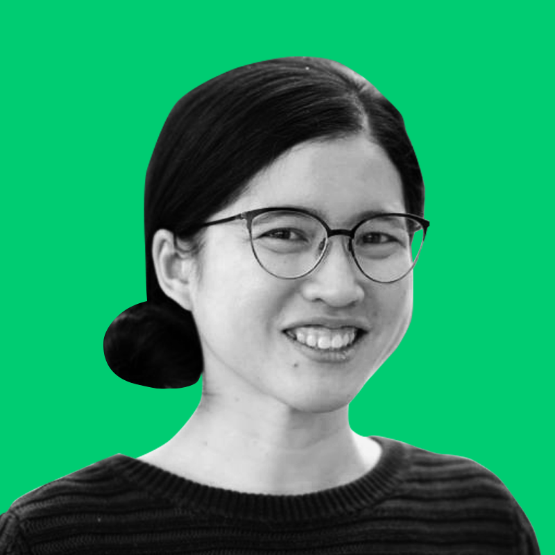 A black and white portrait of a smiling woman with glasses, dark hair tied back, wearing a striped sweater, against a bright green background.