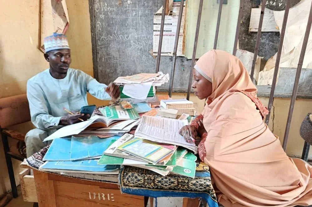 A man and a woman sit at a cluttered desk with various papers and books, in a room with a blackboard in the background.