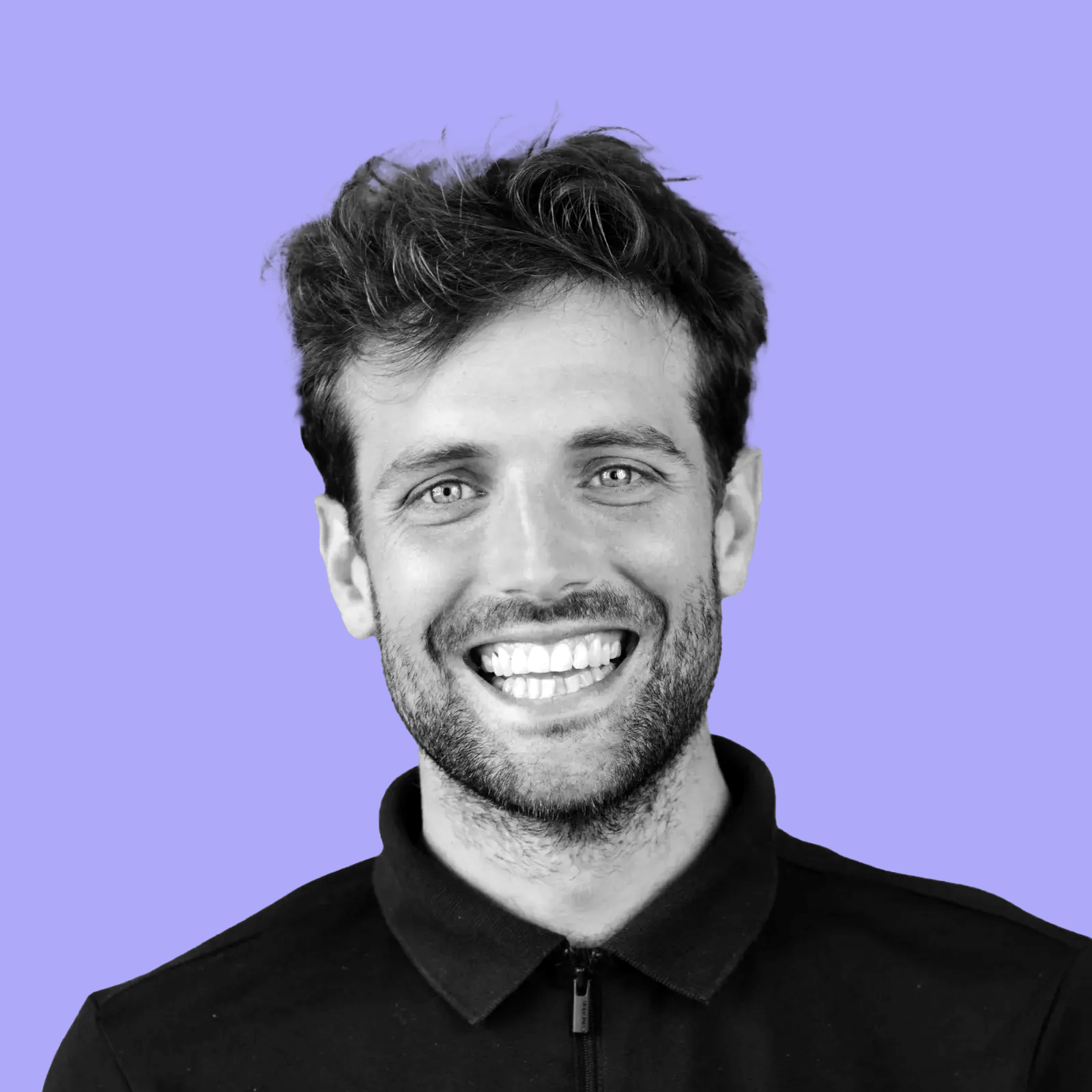 Black and white portrait of a smiling man with light-colored eyes and tousled hair, wearing a black zip-up jacket, against a lavender background.