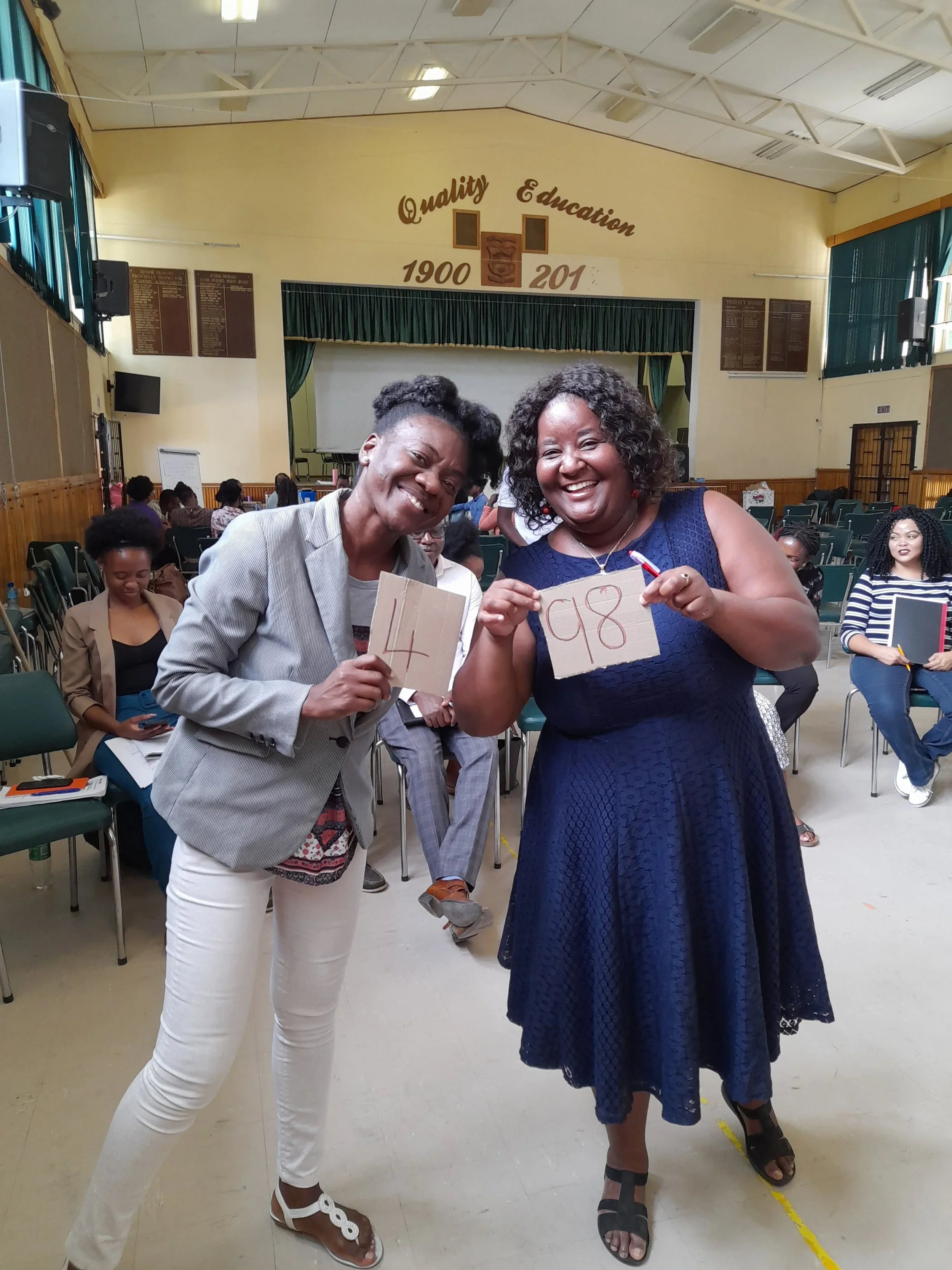 Two women smiling and holding up index cards with the numbers 4 and 98 in a large room with chairs and other people seated in the background.