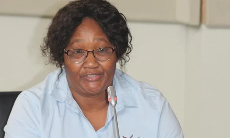 A woman with glasses and curly hair speaking into a microphone during a meeting.