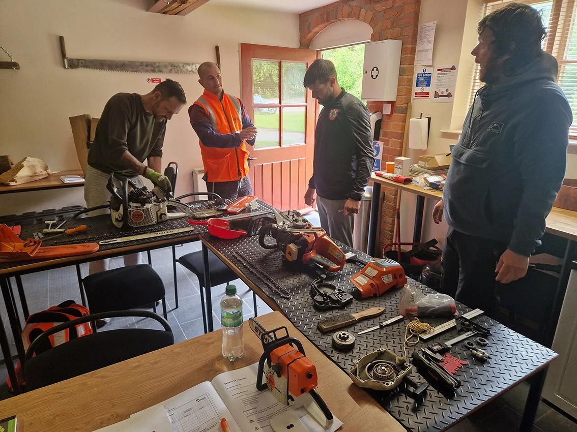 Four men inspecting and working with tools and equipment on tables in a workshop or training room. Items include chainsaws, drills, and safety gear.