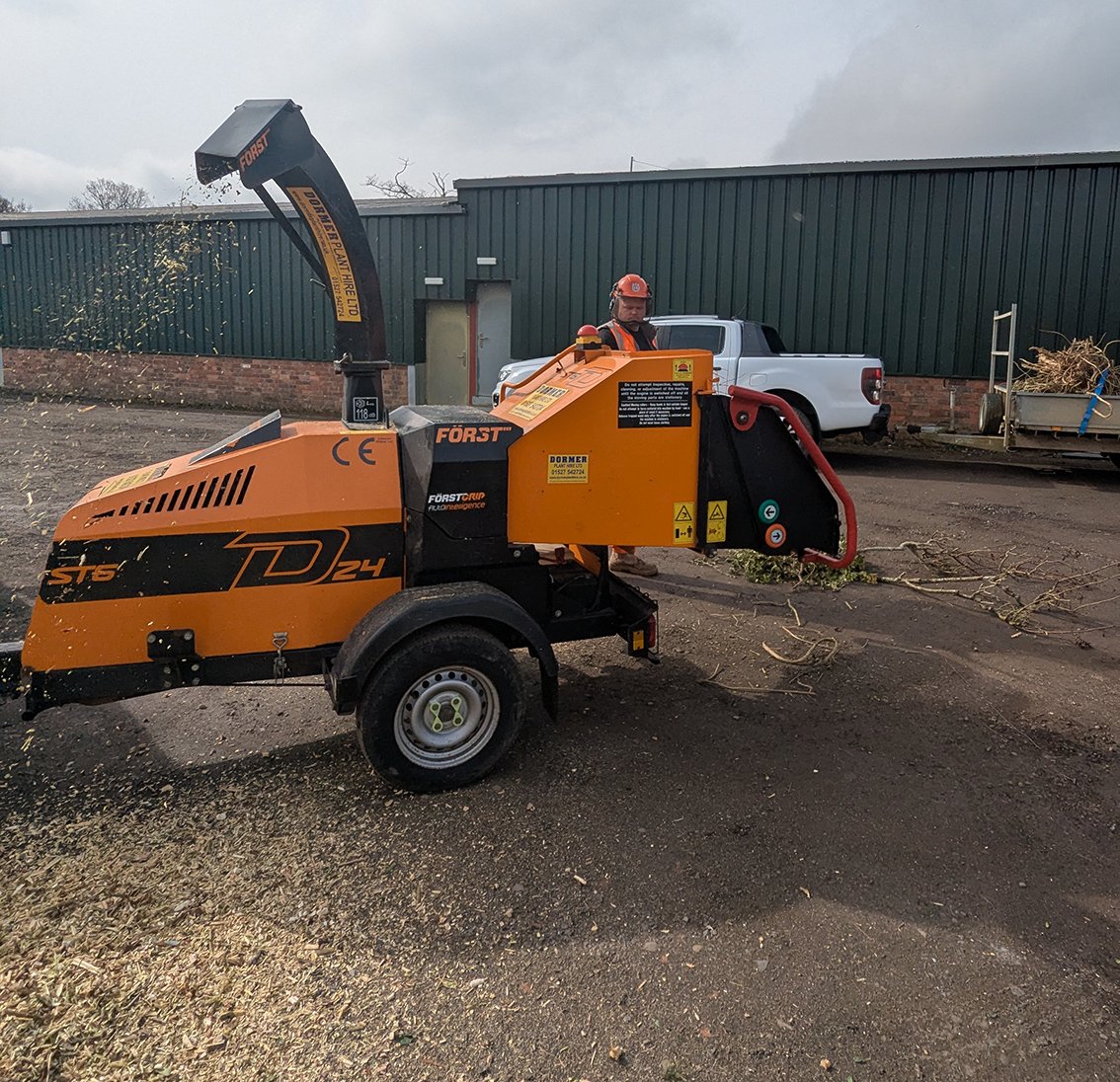 A worker operating an orange wood chipper in an outdoor work site. The worker is wearing safety gear including a helmet and vest.