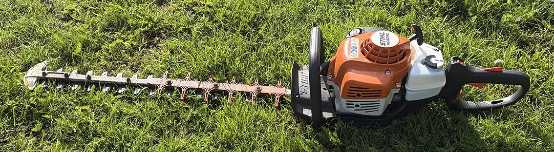 A gas-powered hedge trimmer laying on grass, featuring an orange and white engine and a long, serrated cutting blade.
