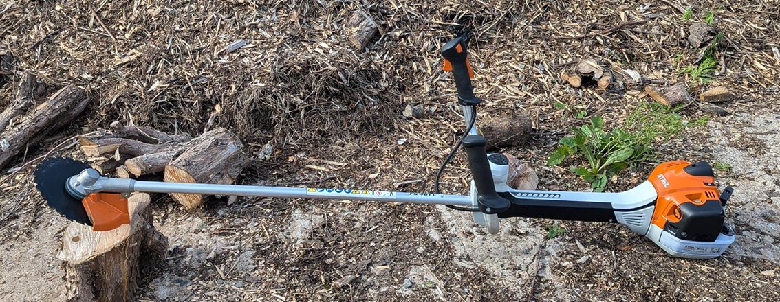 Battery-powered weed trimmer lying on dirt ground next to some chopped wood and green plants.
