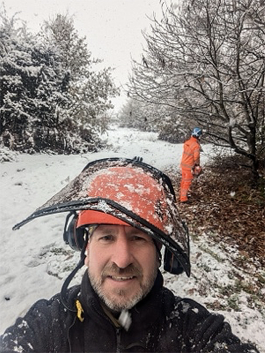 A man in winter clothing, wearing a helmet and sunglasses, takes a selfie on a snowy trail with trees covered in snow, while another person in an orange jacket and helmet walks nearby in a wooded area.