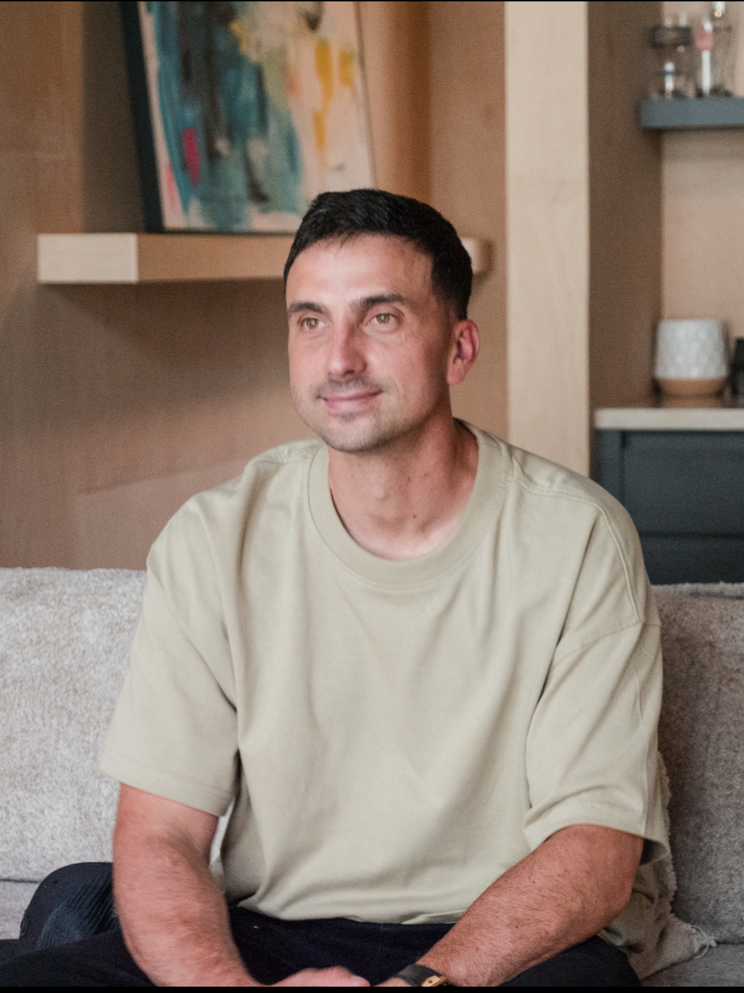 A man with short dark hair and light skin, wearing a beige t-shirt, sitting on a beige couch in a living room with wooden shelves, a colorful abstract painting, and decorative items in the background.