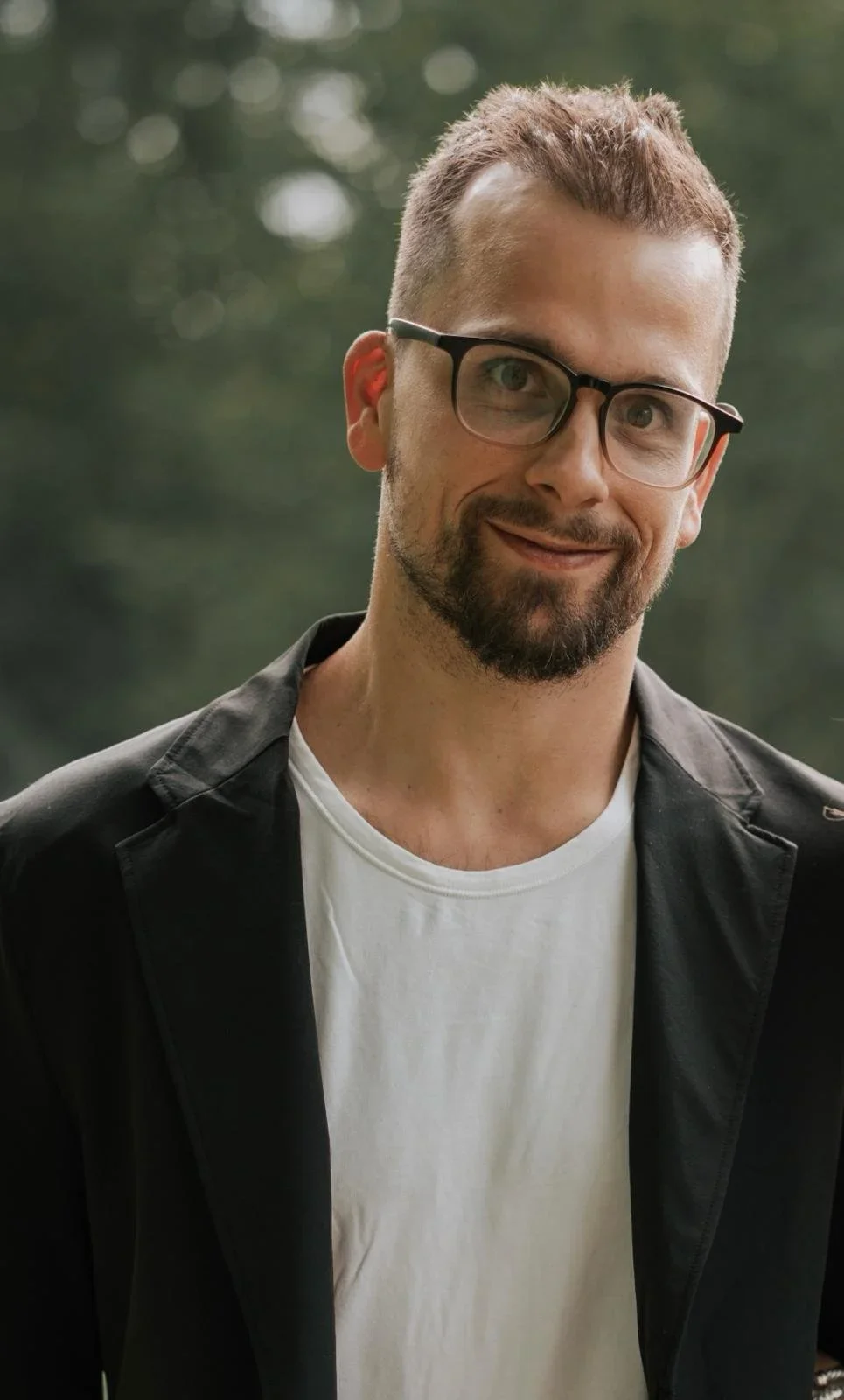 A man with short brown hair, glasses, and a beard wearing a black jacket and white T-shirt, standing outdoors with blurred green trees in the background.