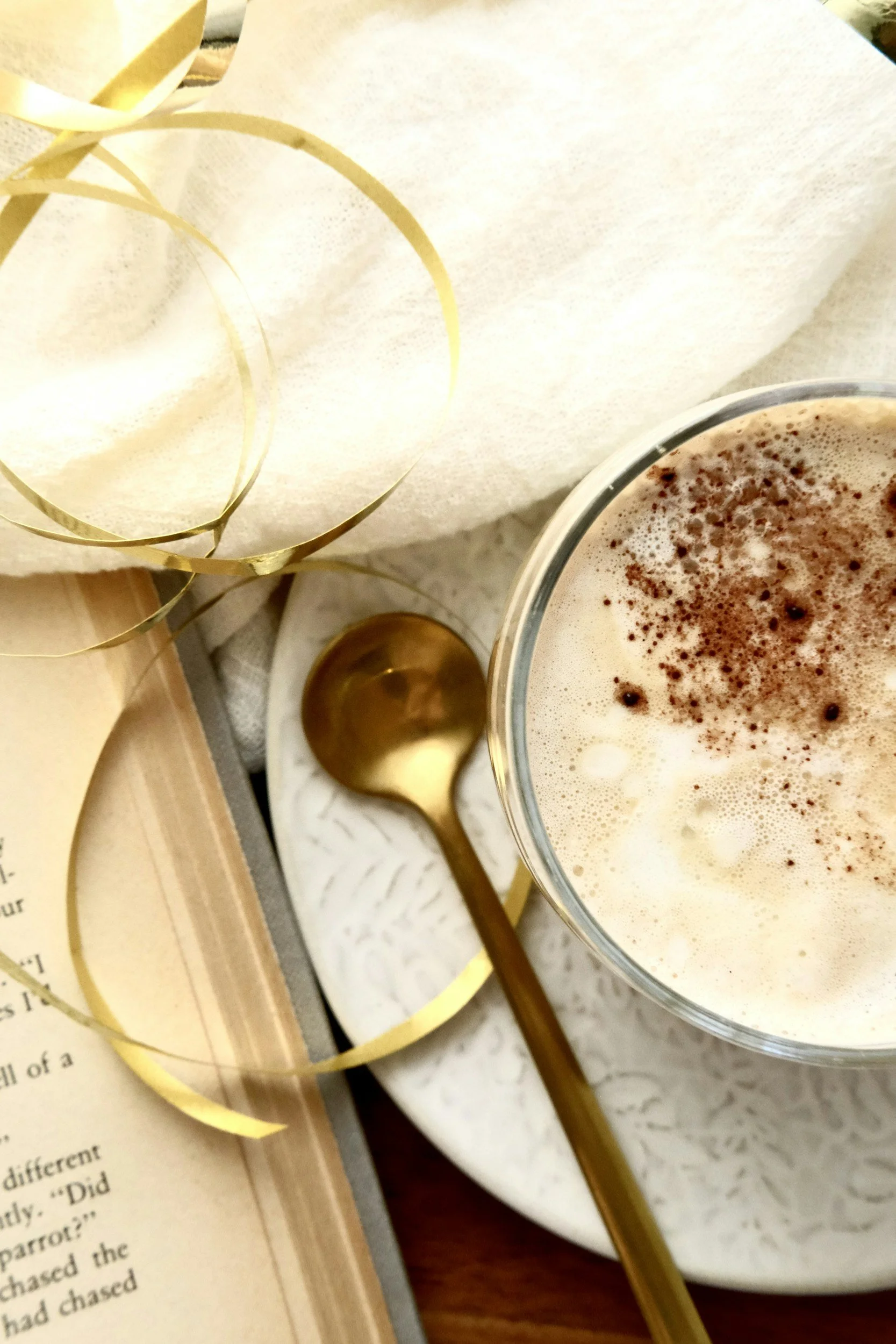 Taza de bebida cremosa con cacao en polvo espolvoreado, cucharada dorada, libro, y decoraciones doradas en un entorno acogedor.