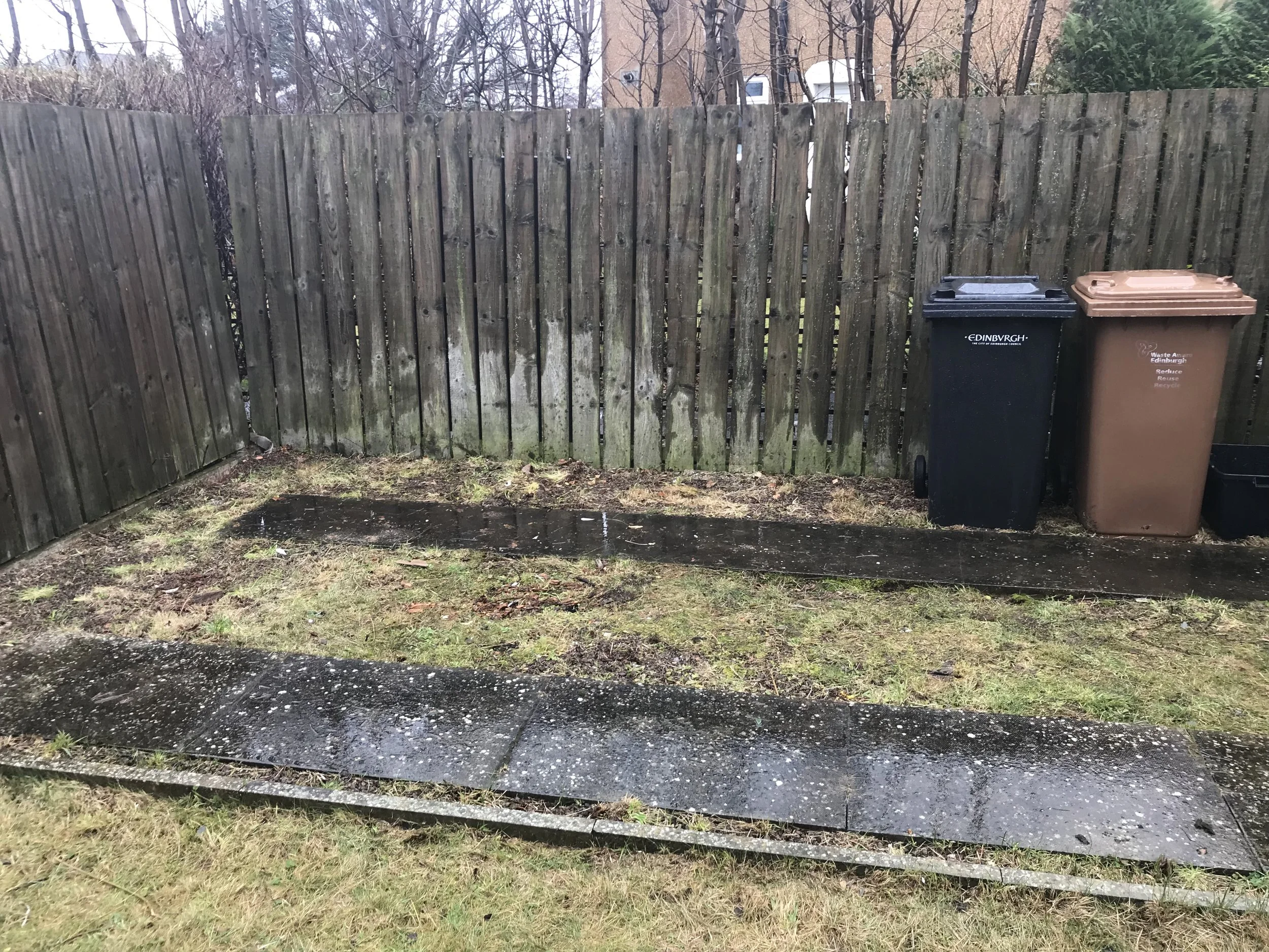 A weathered wooden fence with two waste bins in front, one black and another brown The ground is now cleared from junk, the grass is muddy and wet in front of the fence, with patches of grass and a black tarp-like covering on the ground. 