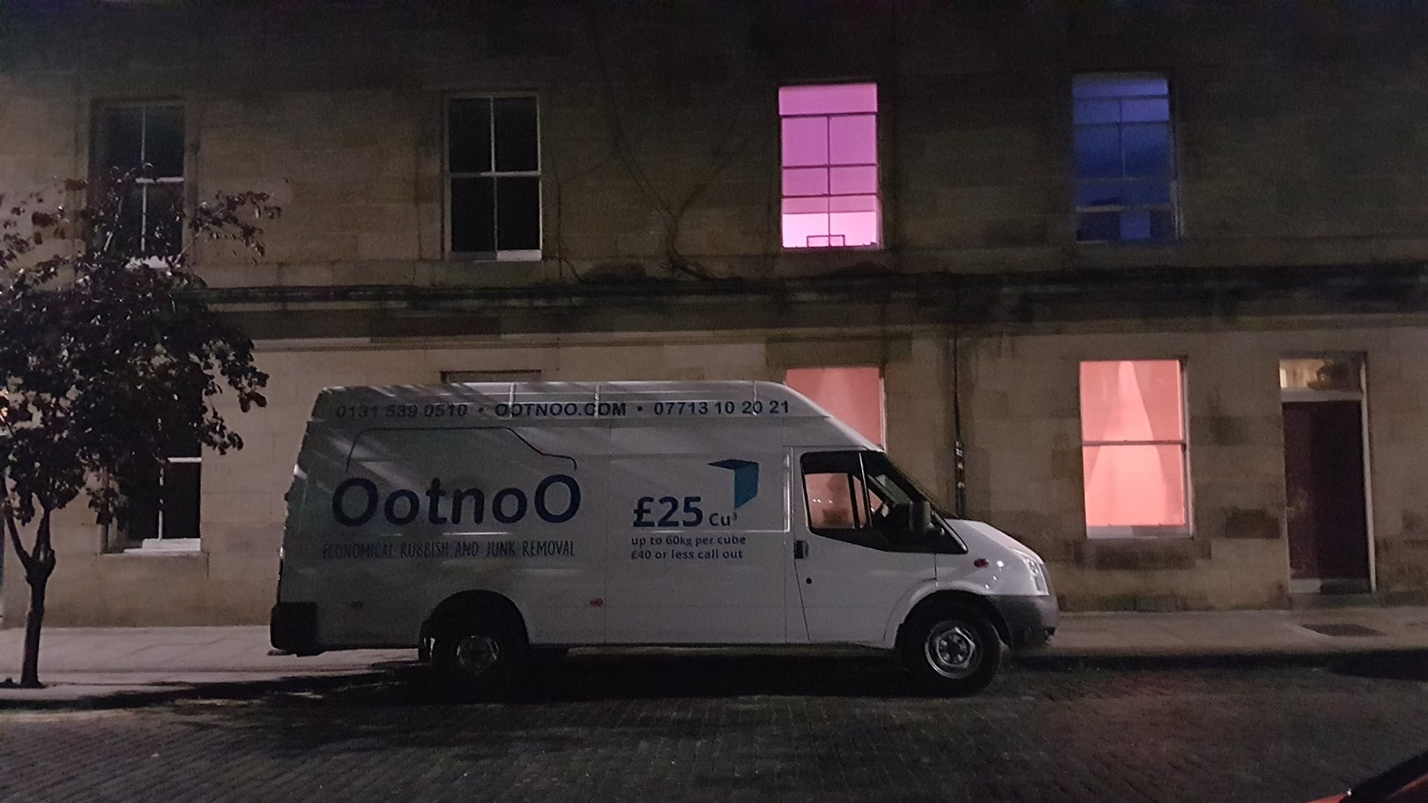 A white van parked on the street at night with advertisement for OotnoO eco-friendly rubbish and junk removal, priced at £25 per cubic detail, with a building in the background featuring lit windows with pink and blue lighting.
