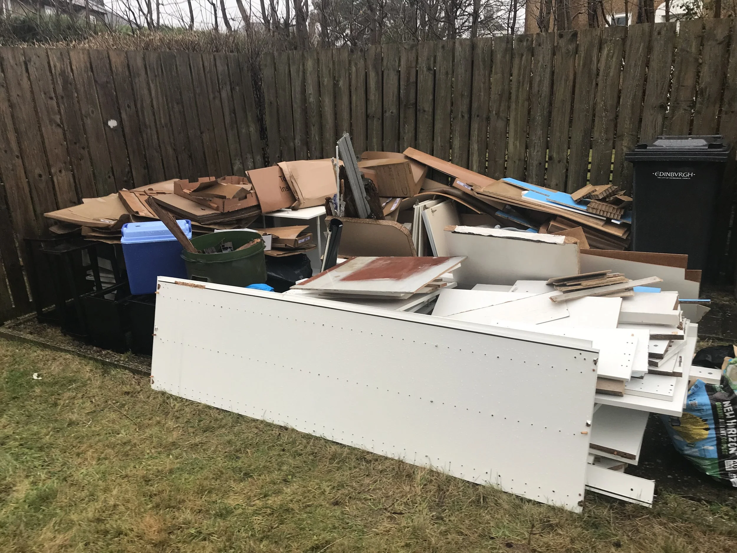 A pile of discarded furniture and household items, including cardboard boxes, furniture pieces, and a trash bin, set against a wooden fence on a grassy yard.