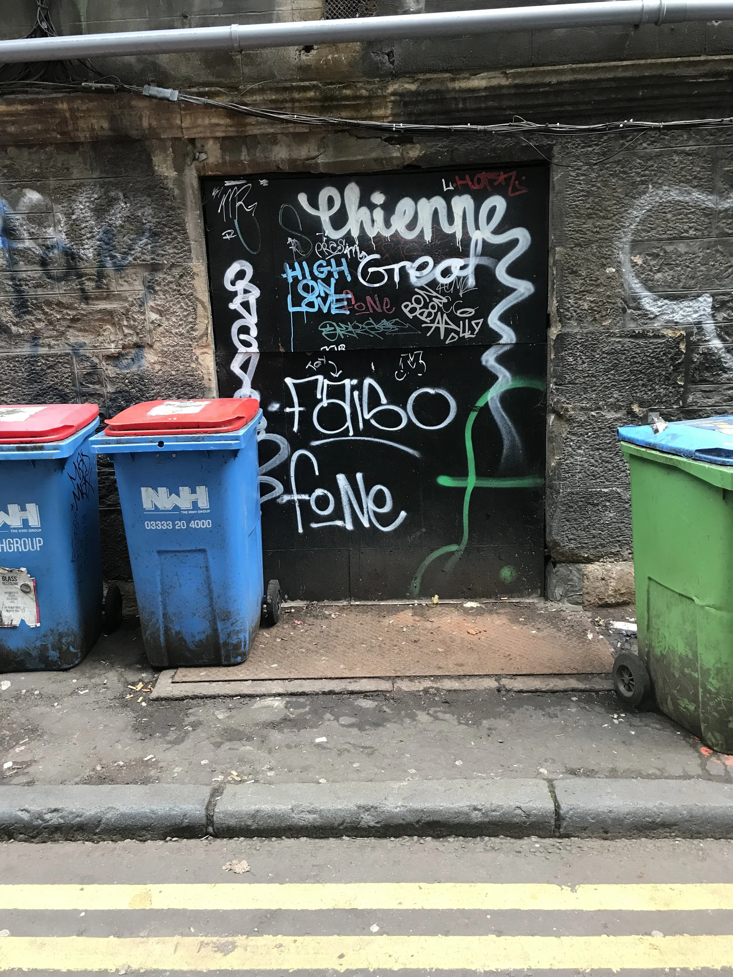 Three waste bins (two blue and one green) are placed on a sidewalk in front of a wall with graffiti. The bags and junk have been cleared, leaving only the commercial bins.  There are also concrete curbs and street markings visible in the foreground.