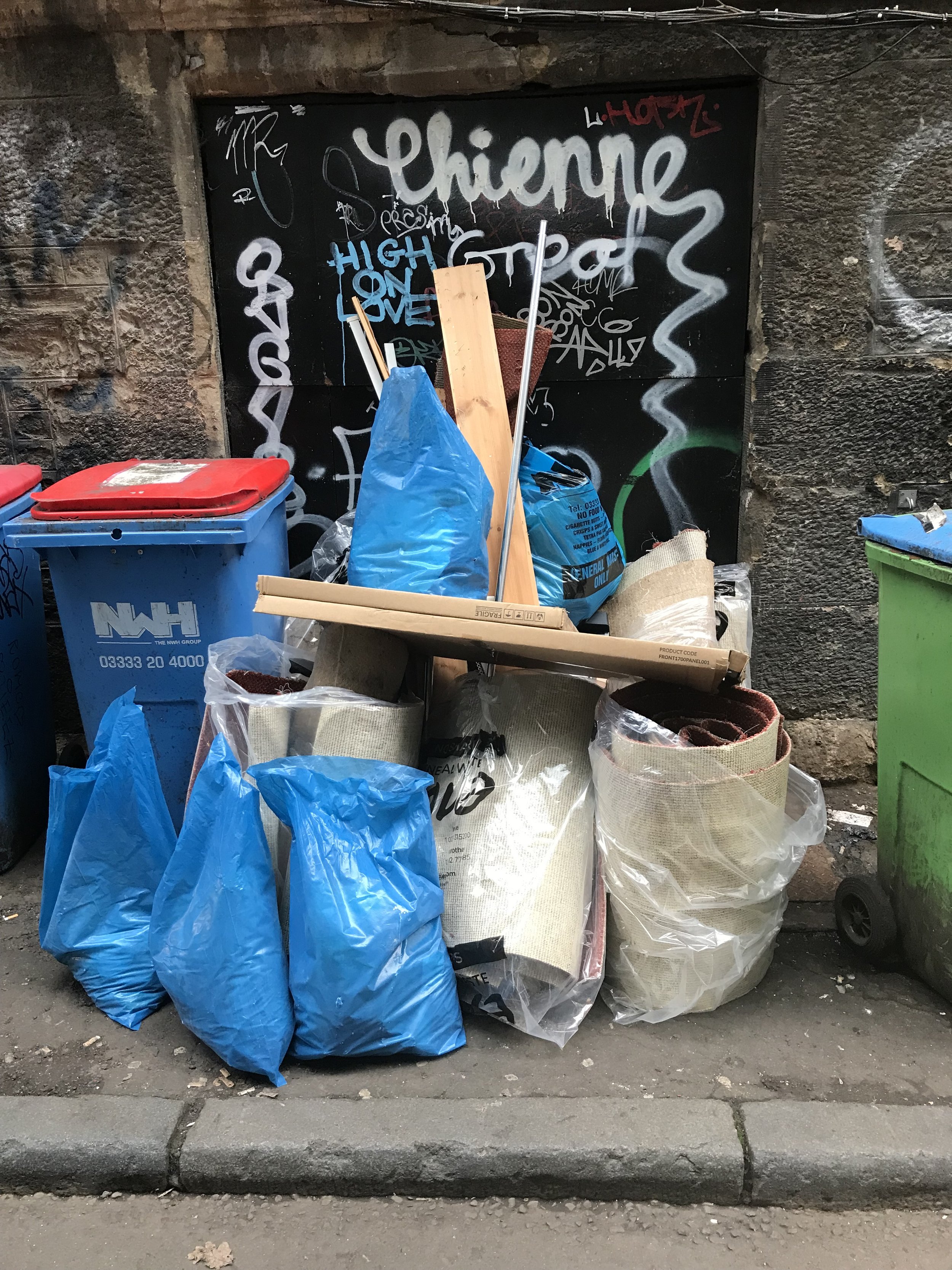 A pile of trash bags, mostly blue, with some white, a wooden plank, a metal pole, and other discarded items, in front of a graffiti-covered wall.
