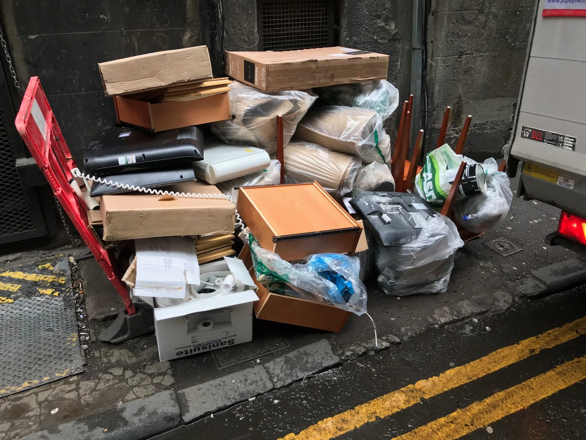 A back street with a pile of discarded furniture, packaging, and electronic items next to a wall with a utility meter. Items include cardboard boxes, plastic bags filled with rubbish.