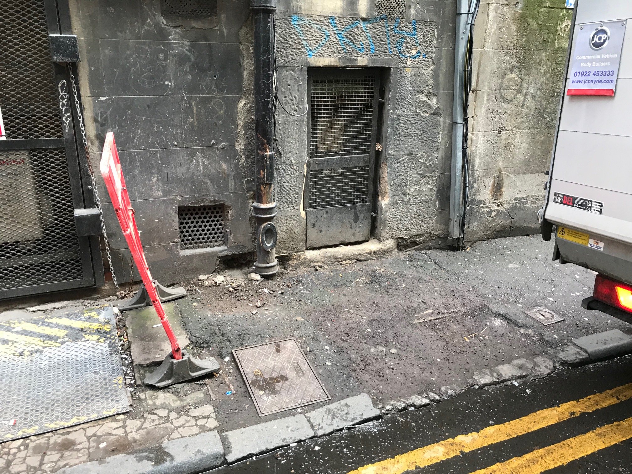 A back street tidied with all the junk cleared, with a clear view of the building wall, a metal gate, a vent, a red ladder, and a utility box. The ground is uneven and dark .