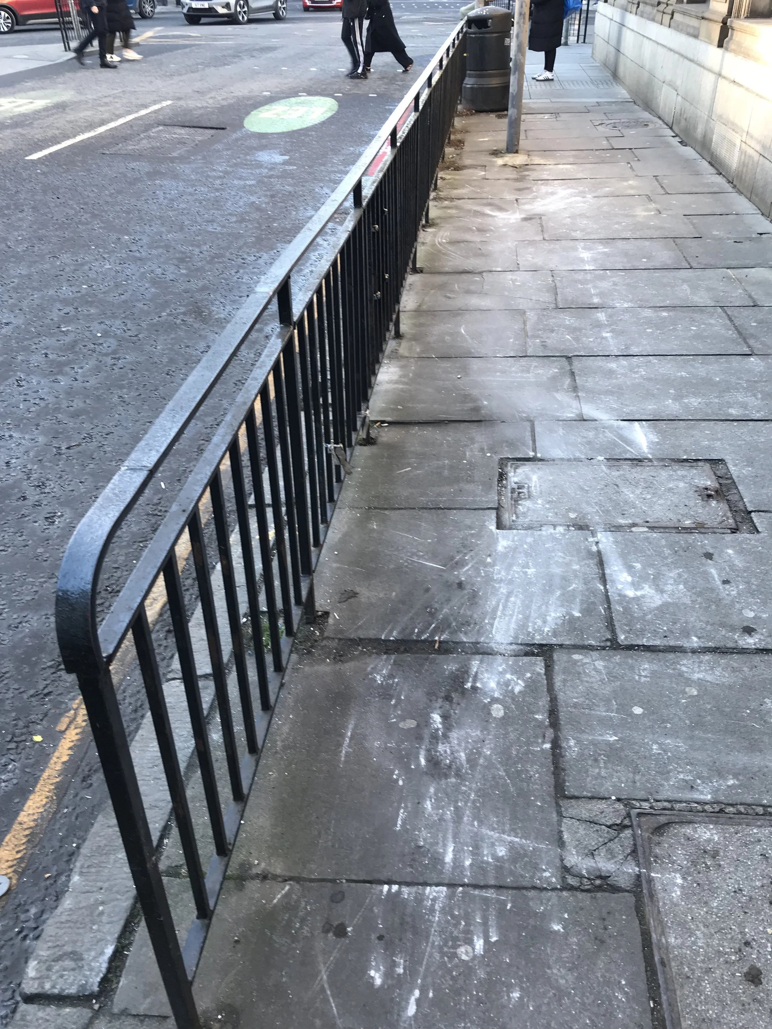 A tidy pavement on a city street with some white chalk markings and a black metal fence separating the path from the road. There are a few pedestrians walking by, and others standing on the pavement.