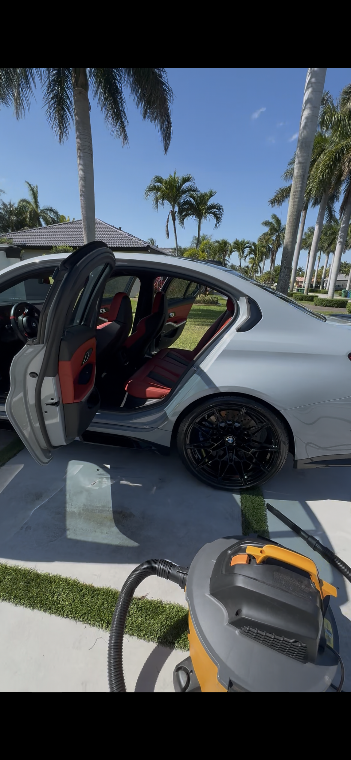 A silver luxury car with red interior seats, parked on a driveway surrounded by palm trees under a blue sky, with a vacuum cleaner in the foreground.
