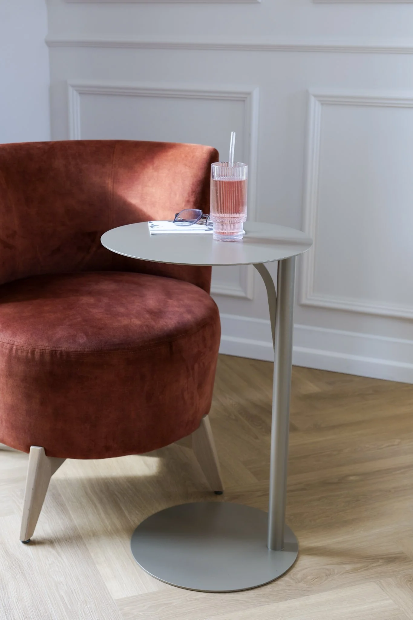 Chaise orange avec un petit livre, une paire de lunettes et une grande verre de boisson rose sur une petite table blanche en face d'un mur blanc avec moulures.