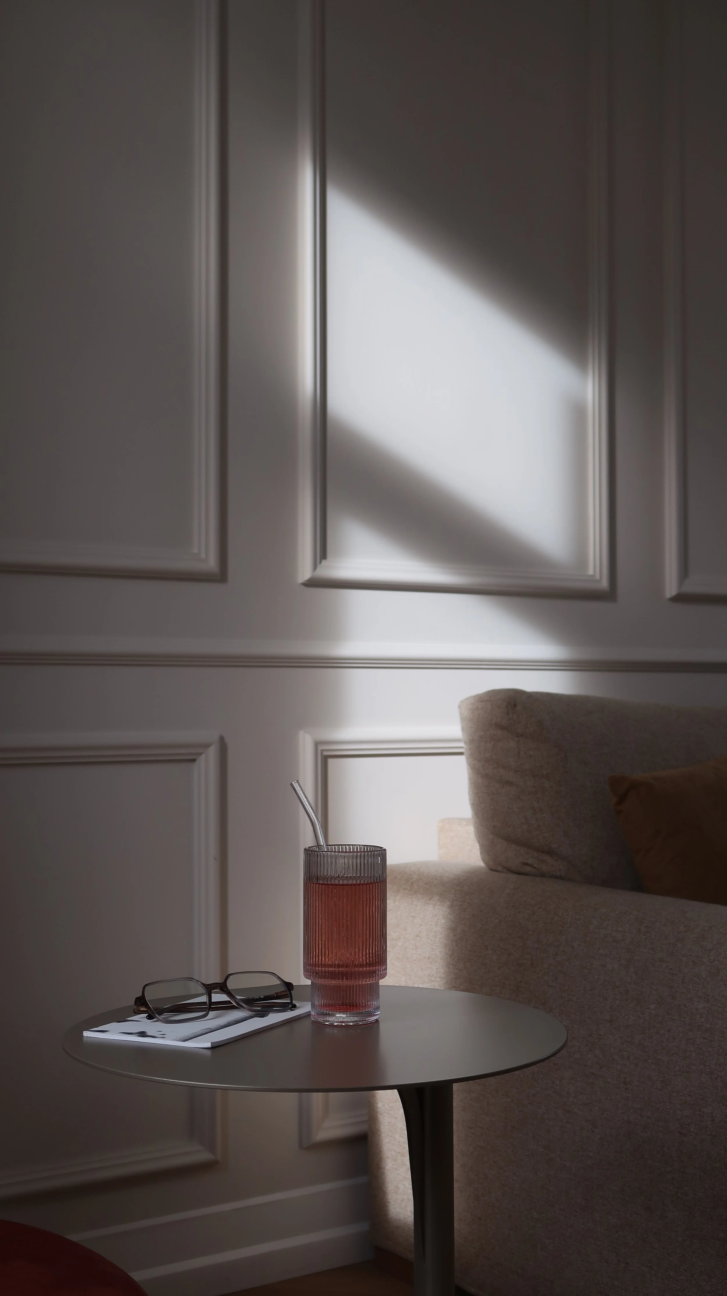 Une table ronde avec une paire de lunettes, un verre à boissons avec une paille, un magazine, une sofa beige avec un coussin marron et un mur blanc avec des moulures, sous la lumière naturelle.