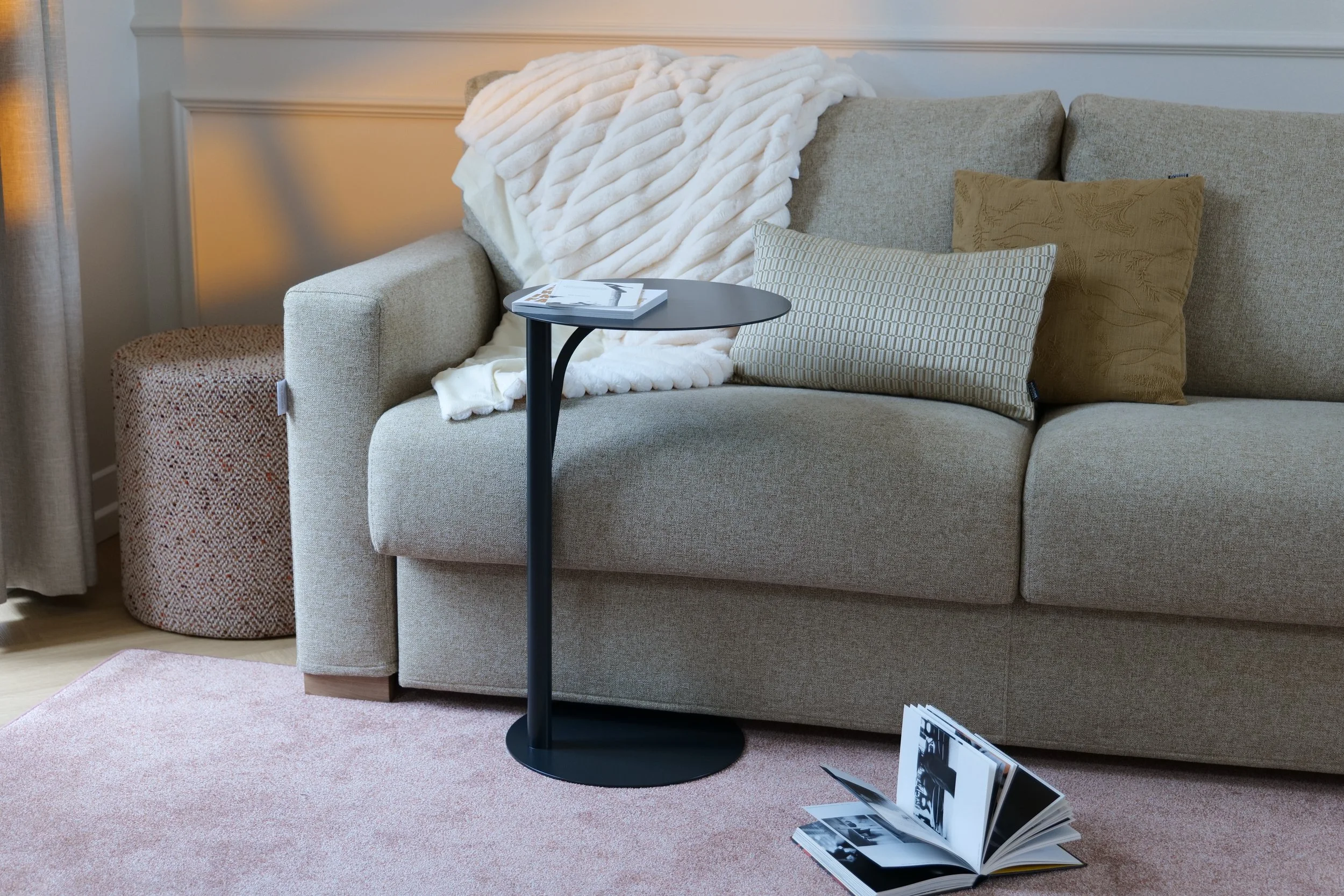Un canapé beige avec deux coussins, une couverture blanche en peluche sur le dossier, une petite table noire avec un magazine dessus, un tapis rose clair, un roman ouvert à terre, et un coin de rideau beige au côté.
