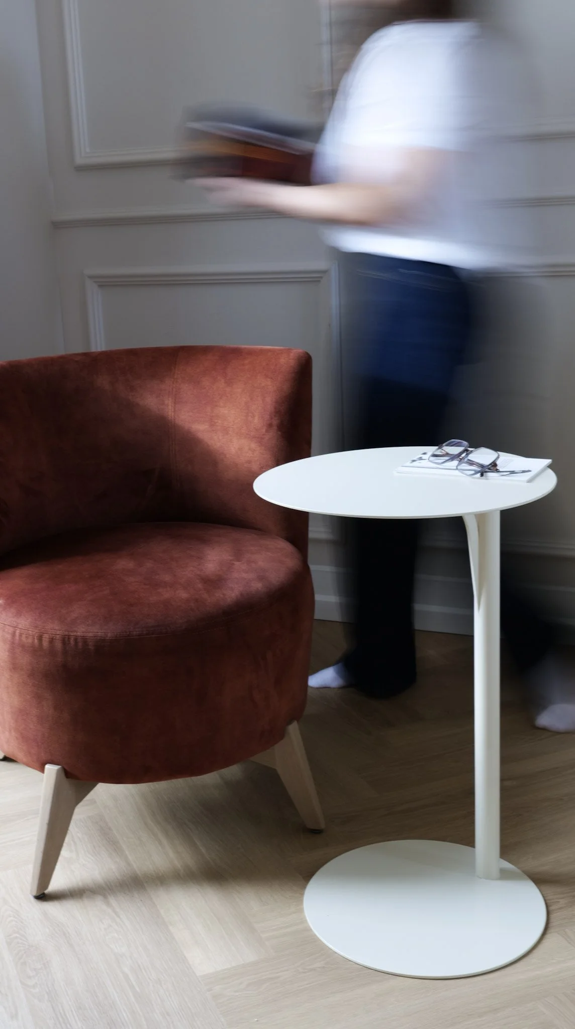 Une personne en mouvement dans une pièce, à côté d’un fauteuil rouge et d’une petite table blanche avec des lunettes et des papiers dessus.