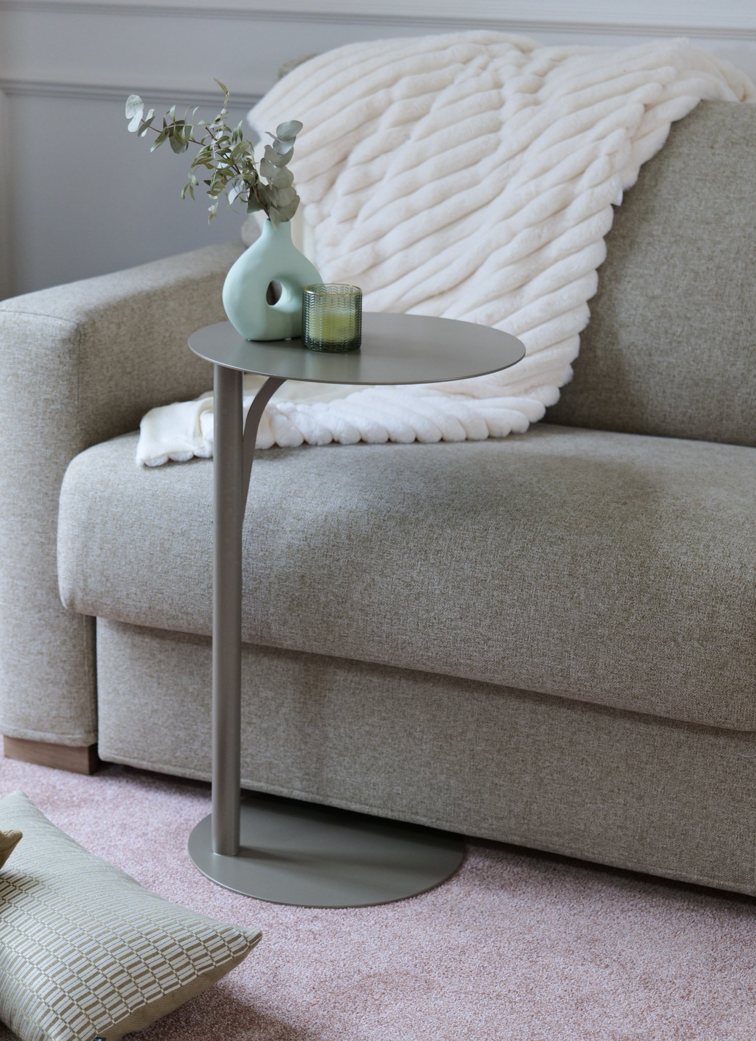 Un canapé beige avec une couverture blanche en peluche, un petit meuble gris avec une vase verte contenant des branches de eucalyptus, et un verre vert. Un coussin beige à carreaux est sur le tapis rose à motifs.