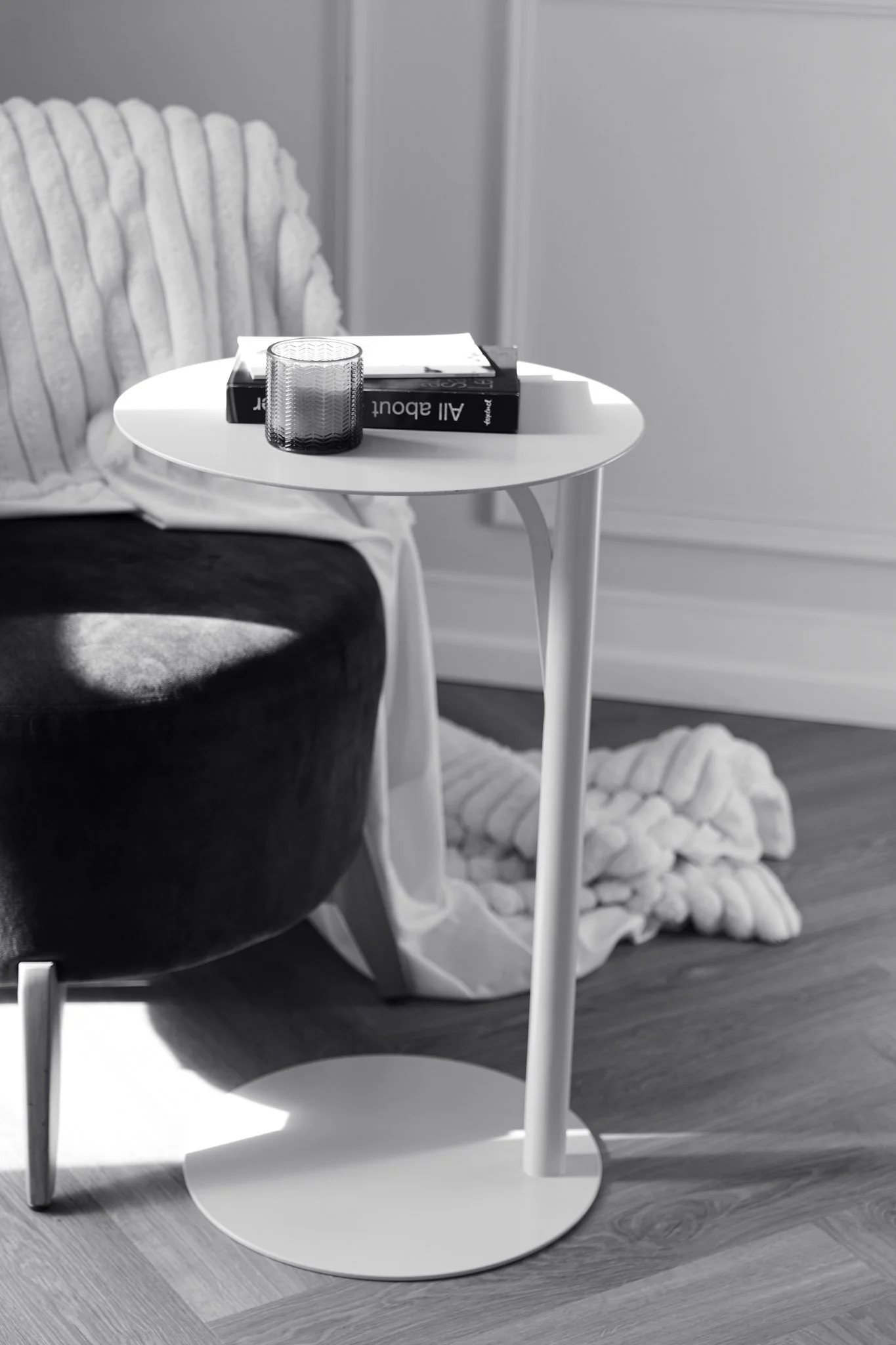 Une table de chevet blanche avec un verre texturé, un livre intitulé 'All About' et un carnet. En arrière-plan, un fauteuil noir et un tapis blanc en peluche sur un sol en bois. La pièce a un style moderne et minimaliste.