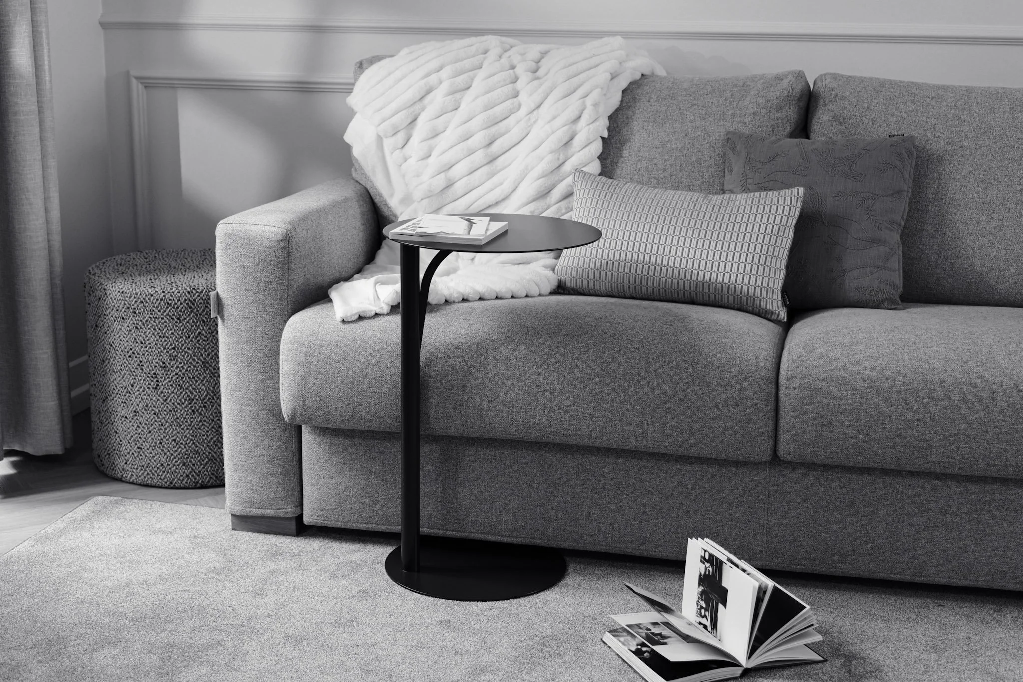 Un salon avec un canapé gris, un coussin rayé et un coussin avec un motif floral, une table noire avec un livre, un plaid blanc sur le canapé, un objet rond en tissu près du canapé et un magazine ouvert sur le tapis.