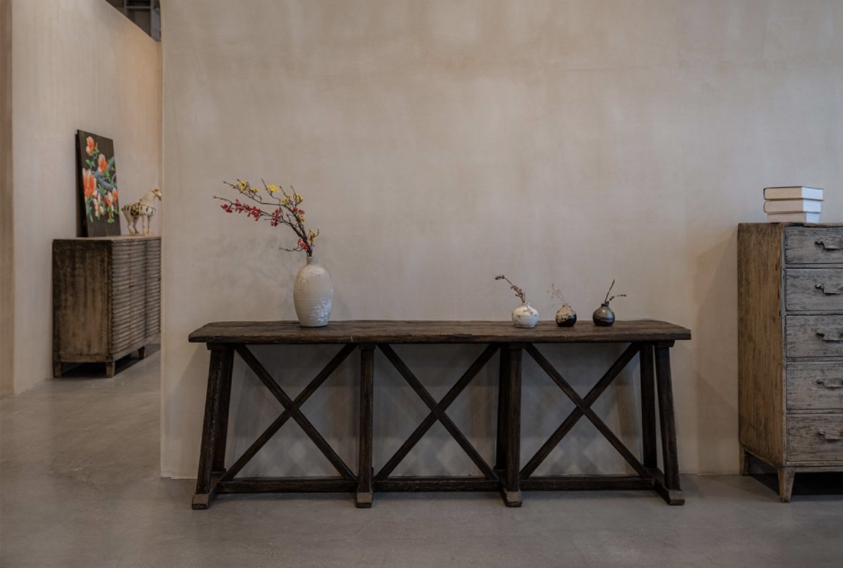 Interior with wooden furniture including a long dark wood table with decorative vases and branches, a light-colored dresser on the right with white books on top, and a cabinet with a flower painting and a decorative animal on the left