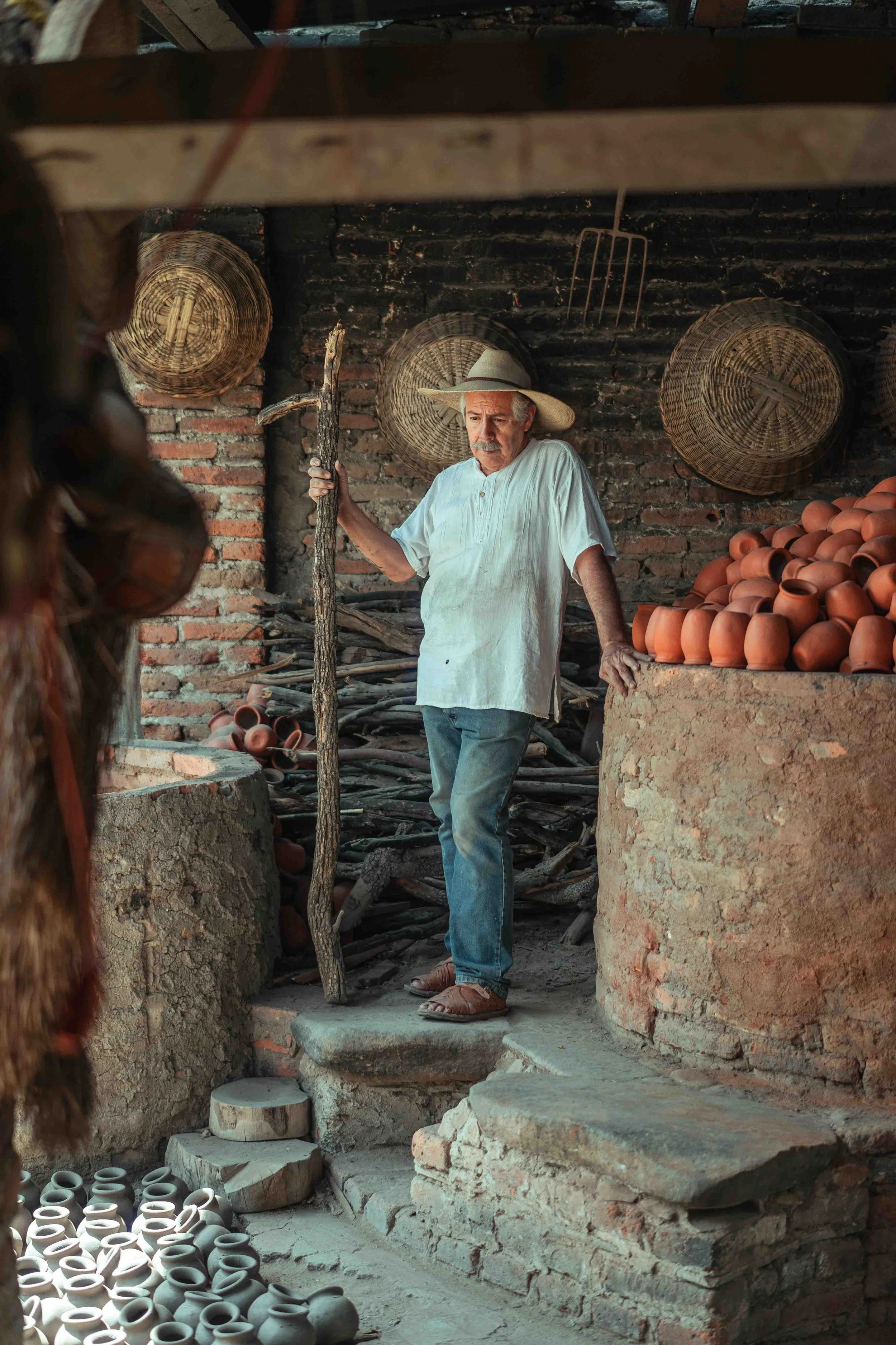 An elderly man with a white shirt, jeans, and a wide-brimmed hat stands on a stone step inside a rustic pottery workshop. He holds a wooden staff in one hand and gazes downward. Behind him are brick walls decorated with woven baskets, and to his right, there is a large pile of small, reddish-brown clay pots.