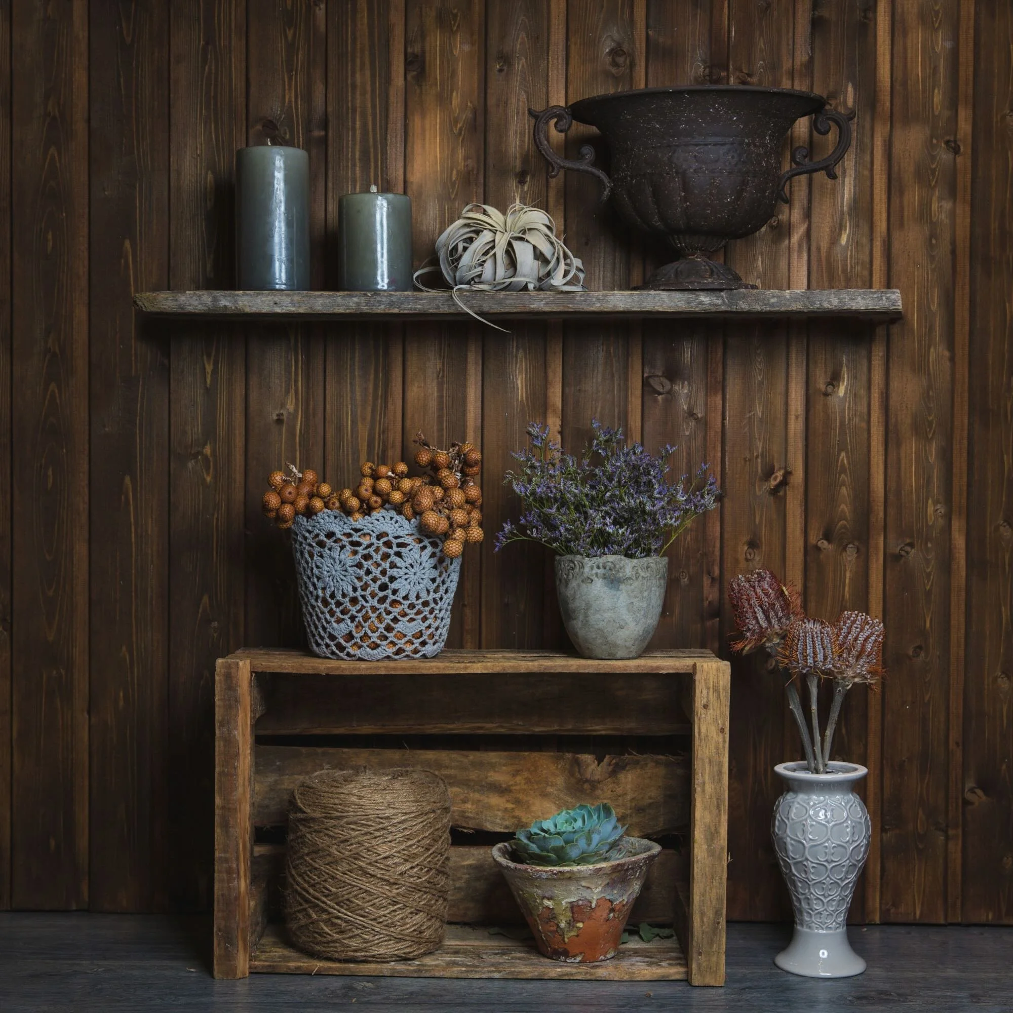 Decorative indoor arrangement with candles, a large container, dried flowers, a basket of berries, a potted plant, a spool of twine, a succulant, and a ceramic vase with dried branches against a wooden wall.
