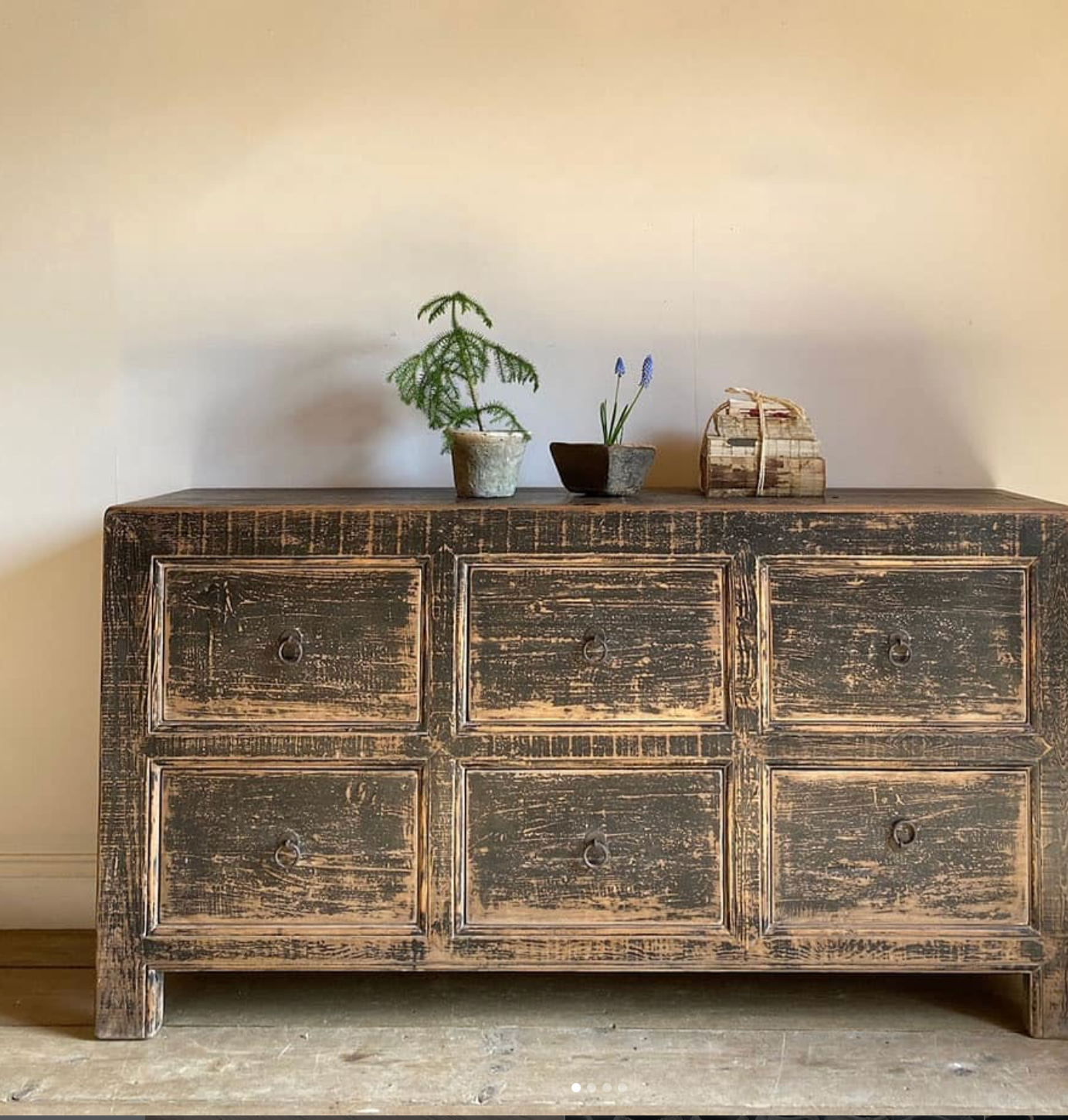 A vintage wooden dresser with six drawers displaying a distressed finish, topped with three small potted plants and a small decorative box.