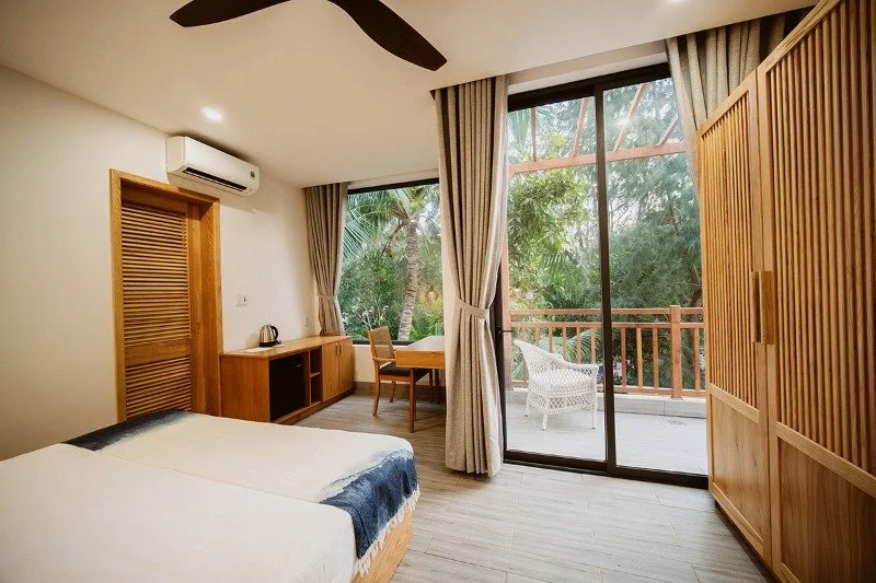 A hotel room with a bed, wooden furniture, large sliding glass door with curtains, an outdoor balcony with a white chair, and lush green trees outside.