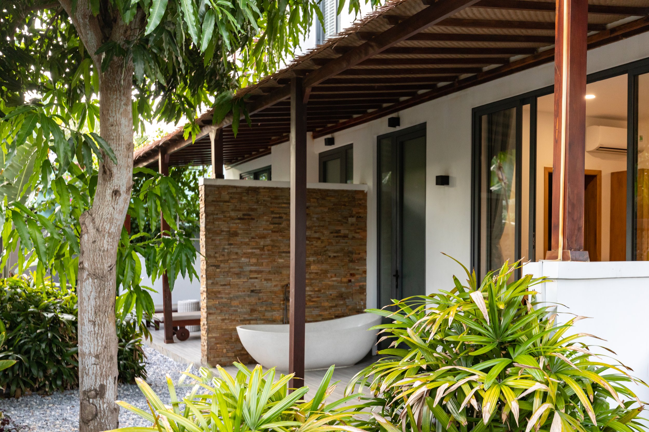 Exterior view of a modern house with a porch, sliding glass doors, and a bathtub outside, surrounded by lush greenery and plants.