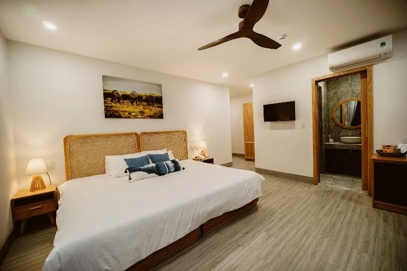 A hotel room with a large bed, nightstands with lamps, a painting above the bed, a wall-mounted TV, a ceiling fan, an air conditioner, and a doorway leading to a bathroom with a mirror and sink.