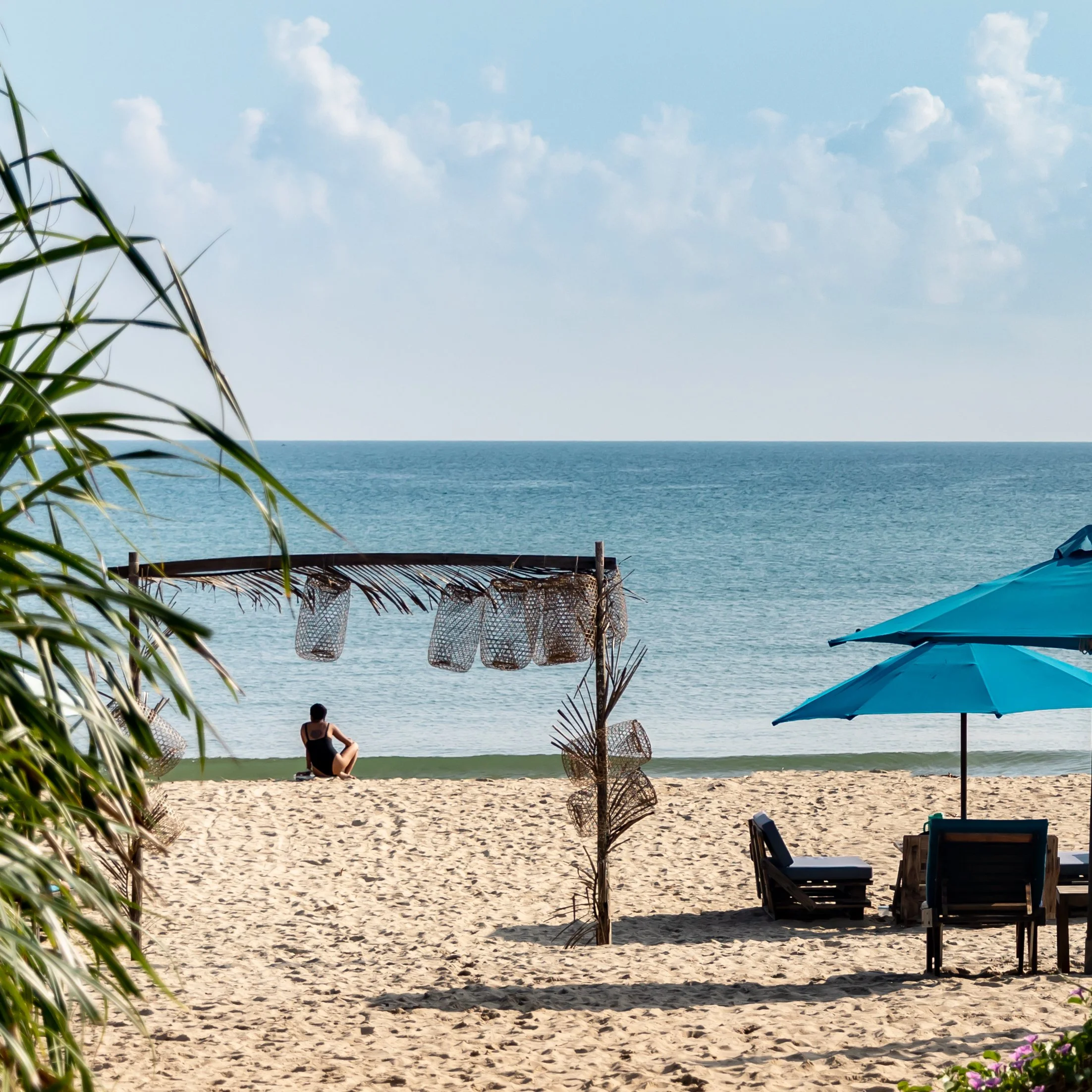 A person sitting on the beach facing the ocean, with a decorated wooden frame and hanging baskets nearby, under a mostly cloudy sky.
