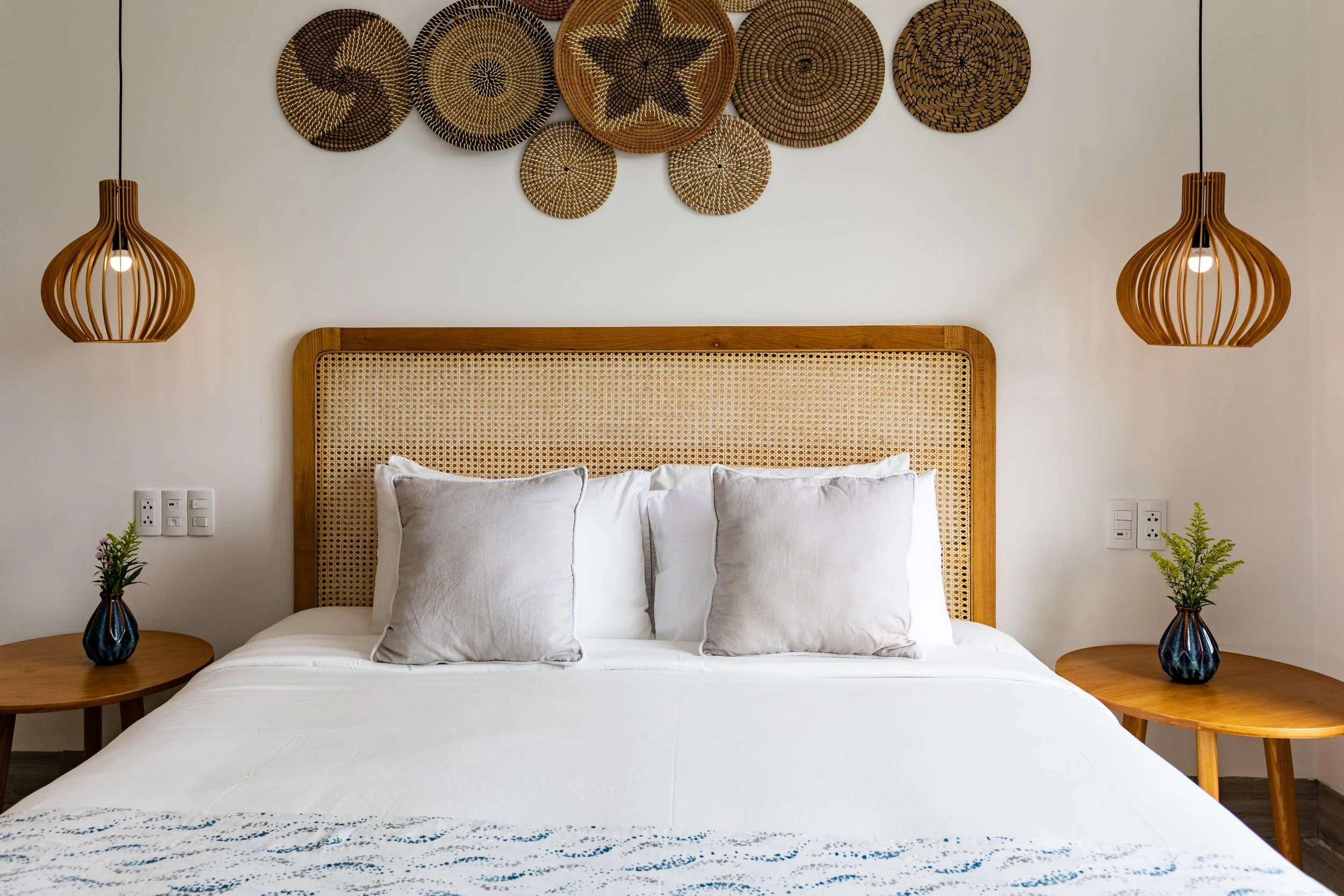A bedroom with a wooden headboard, white bedding, and two pillows. There are two wooden side tables with small plants in blue vases. Two pendant lights hang on either side of the bed. Over the bed, there is wall decor made of woven baskets in various