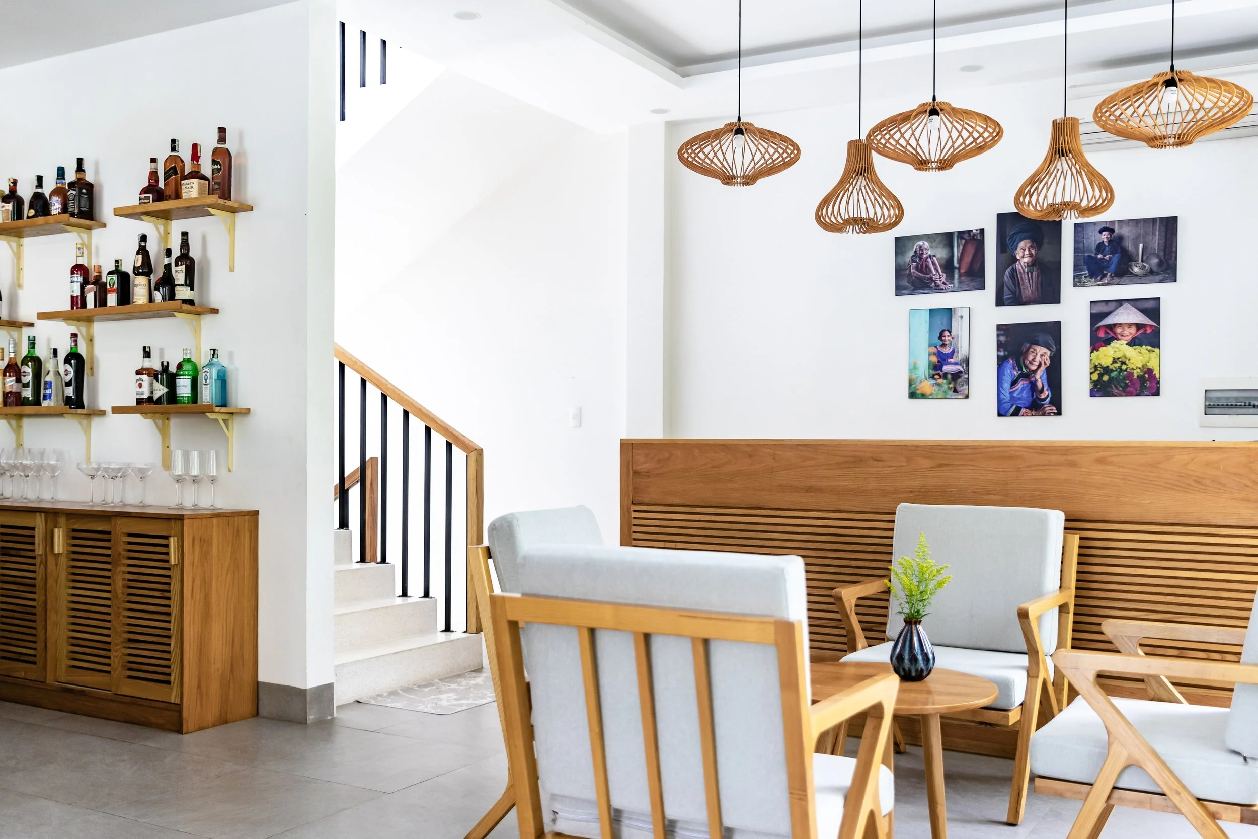 Interior of a modern lounge area with wooden furniture, white cushioned chairs, a wooden table with a small vase, wall-mounted photographs of people, pendant lamps, and a wall-mounted shelf with bottles and glassware.