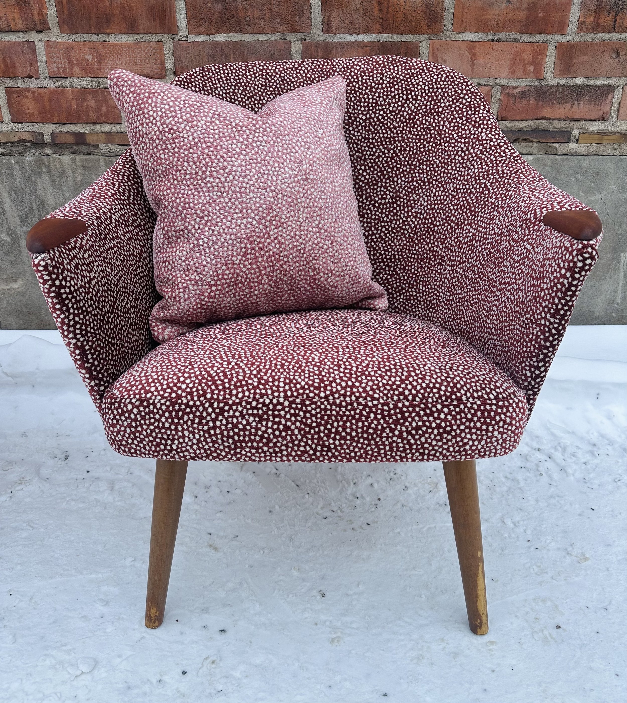 Punainen ja valkoinen kuvioitu nojatuoli ja tyyny, taustalla tiiliseinä ja luminen maa.