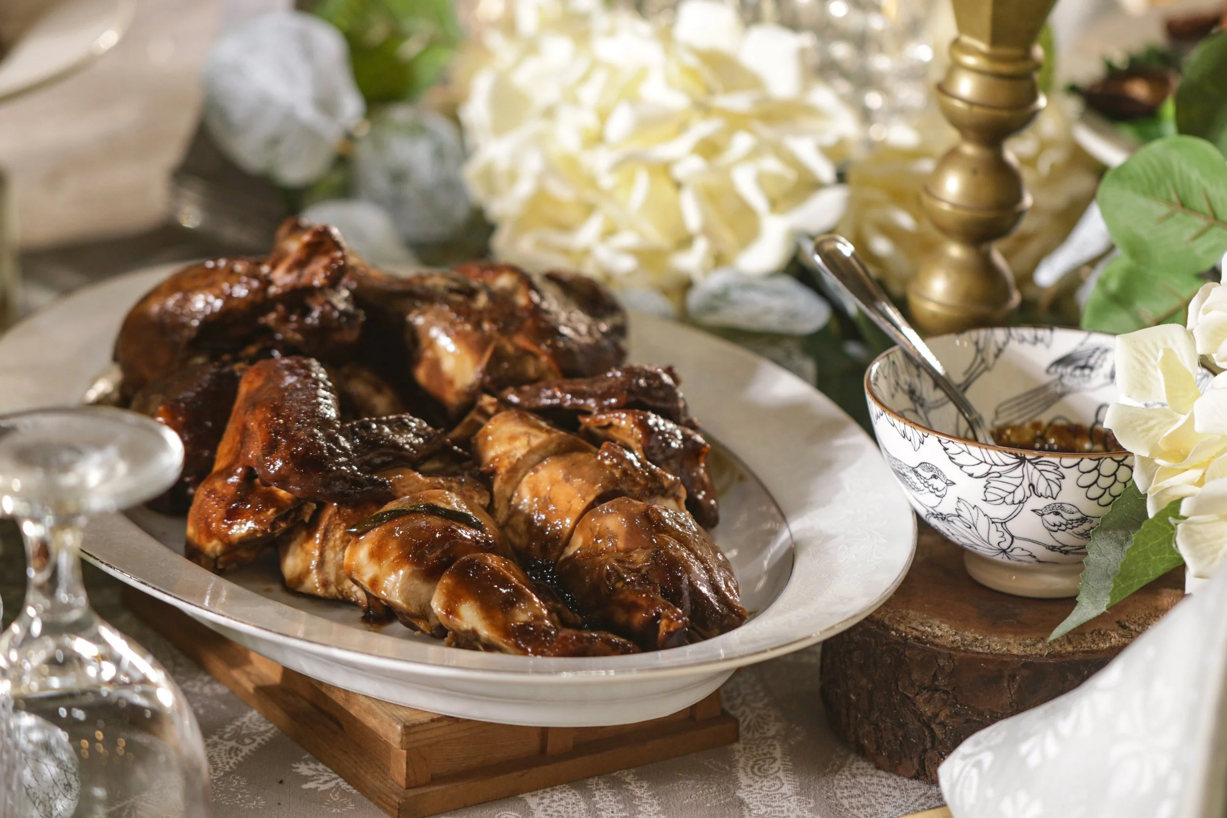 A serving platter with glazed chicken pieces on a wooden base, surrounded by decorative flowers and a bowl of sauce with a spoon, on a textured tablecloth.