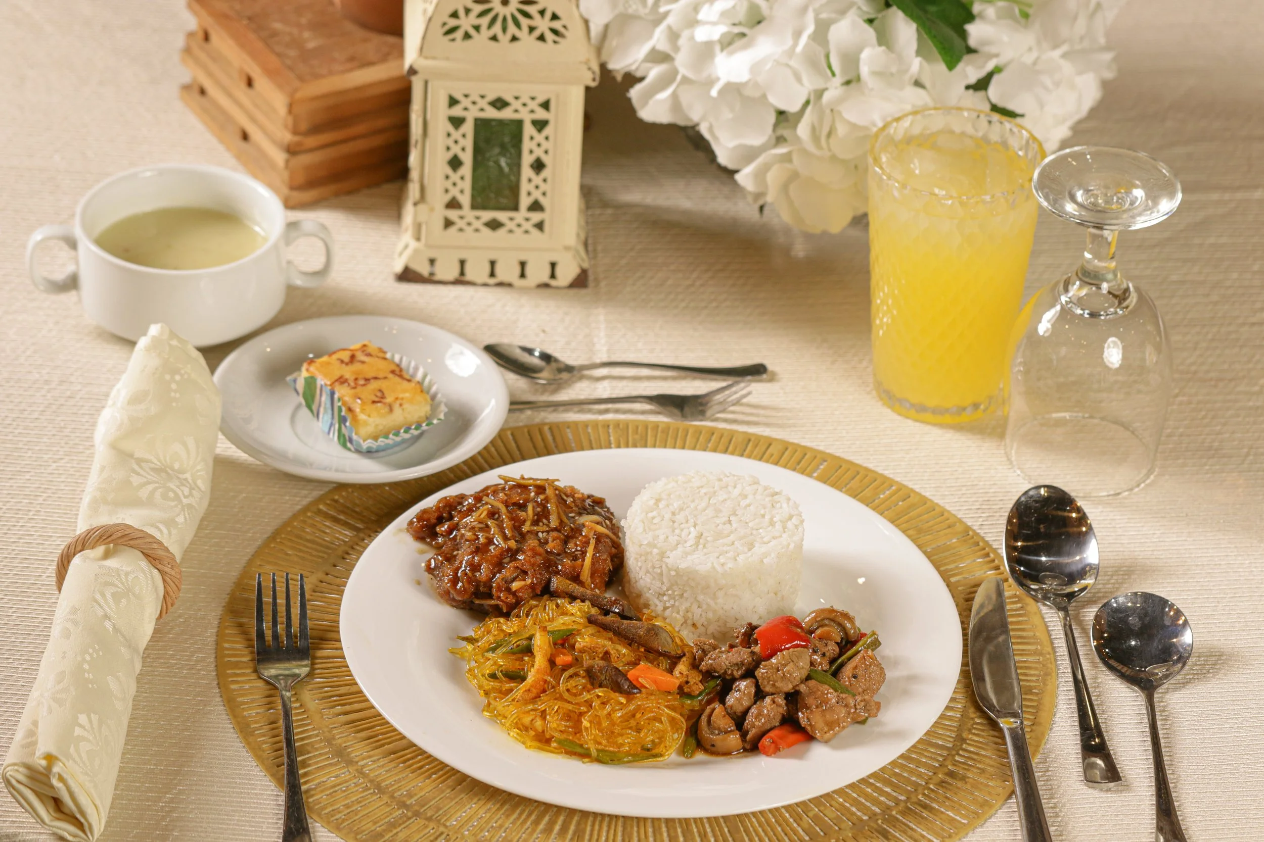 A formal dinner table with a plate of rice, beef with vegetables, noodles, and a stew, accompanied by soup, juice, and a glass of wine or champagne. Silverware and a rolled napkin are arranged, with decorative items and flowers in the background.