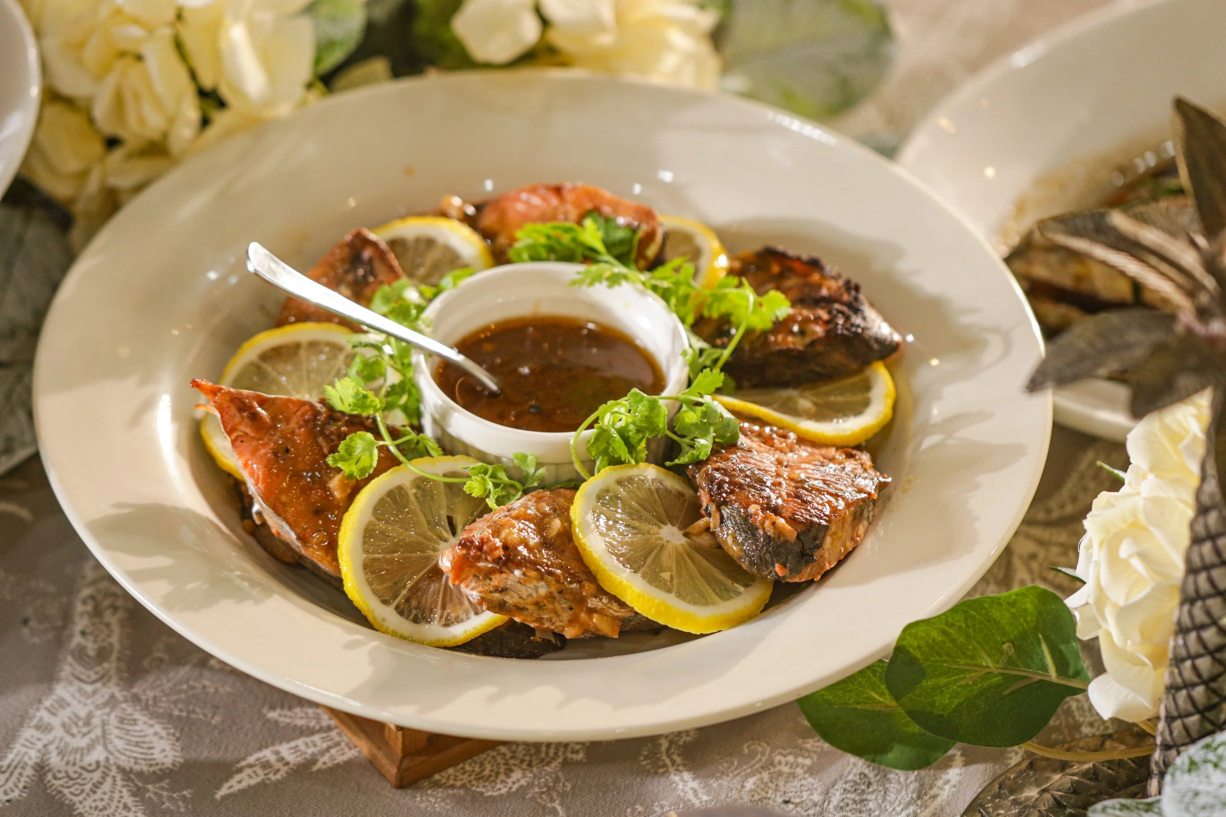 Plate of grilled fish garnished with lemon slices and cilantro, with a side of dark sauce in a small dish, on a decorative table with flowers and leaves.